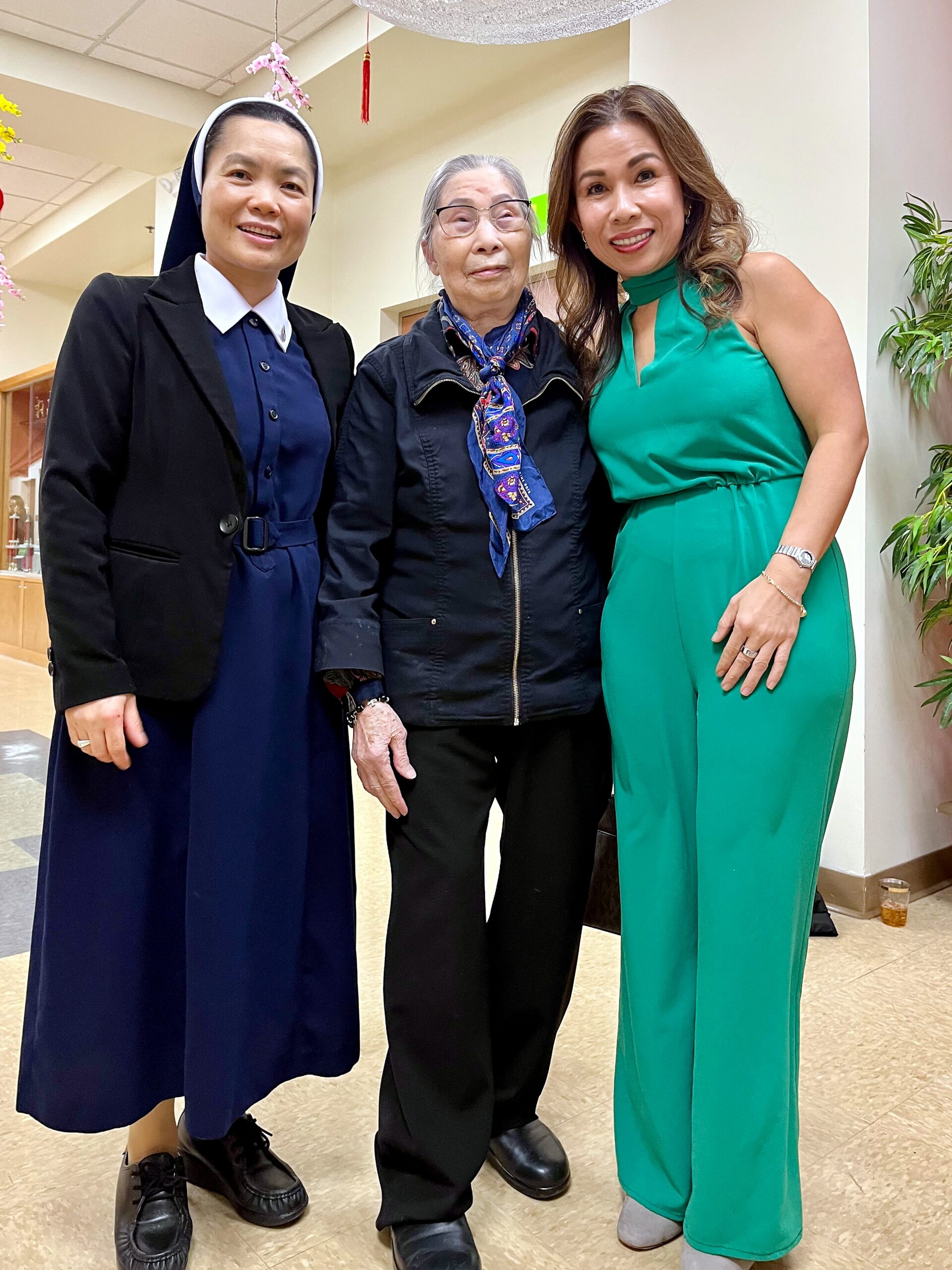 Three women posing indoors and smiling at the camera: left in a dark blue dress with blazer and head covering, middle an older woman in a black jacket with a blue scarf, right in a bright green jumpsuit.
