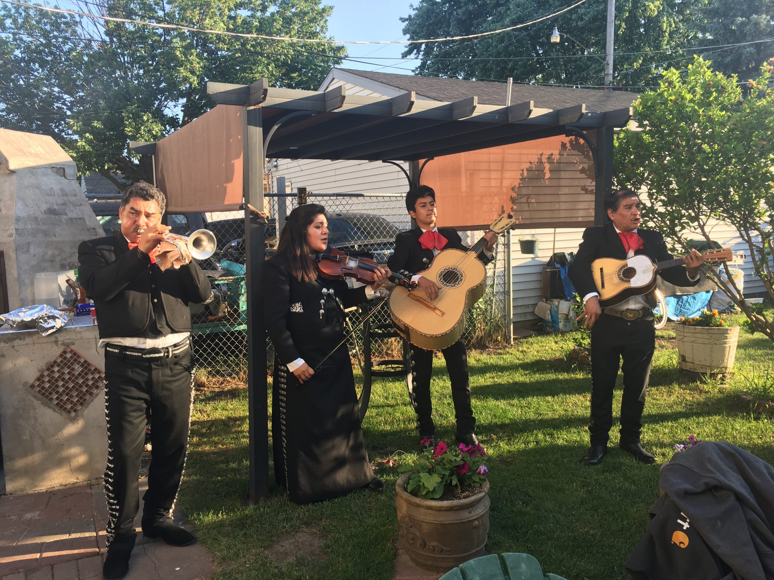 Mariachi quartet performing outdoors in a backyard, four musicians in black charro outfits with red bow ties, playing trumpet, violin, and guitars under a patio cover.
