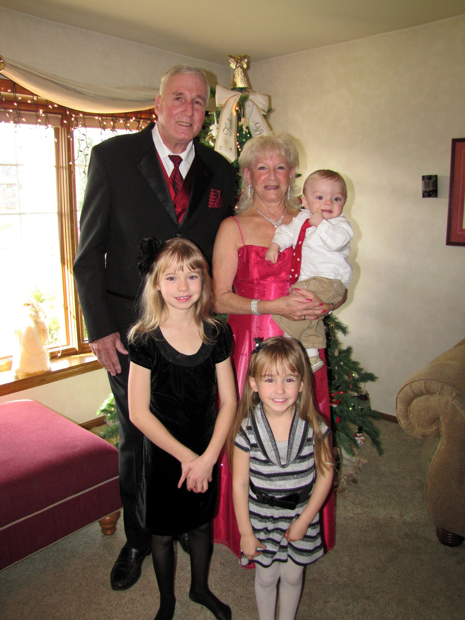 Multigenerational family posing for a holiday photo in a living room, with a Christmas tree in the background; grandparents in formal wear and three children in festive outfits.