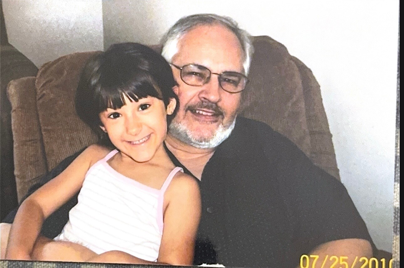 Grandfather and granddaughter sitting together on a brown recliner, both smiling at the camera.