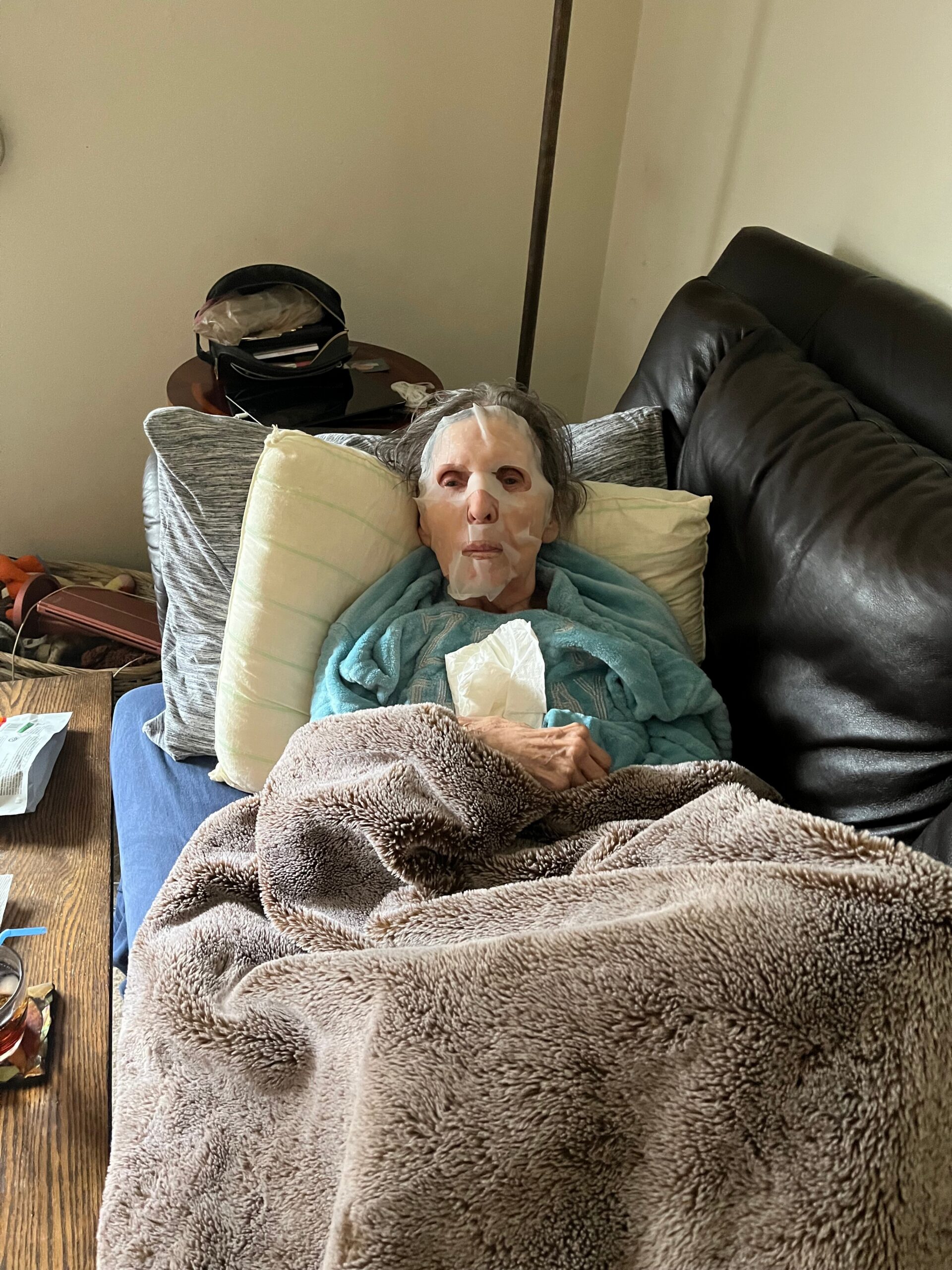 Person lying on a blue blanket on a sofa wearing a white facial mask, resting with pillows and a brown fuzzy blanket. A side table and clutter are in the background.