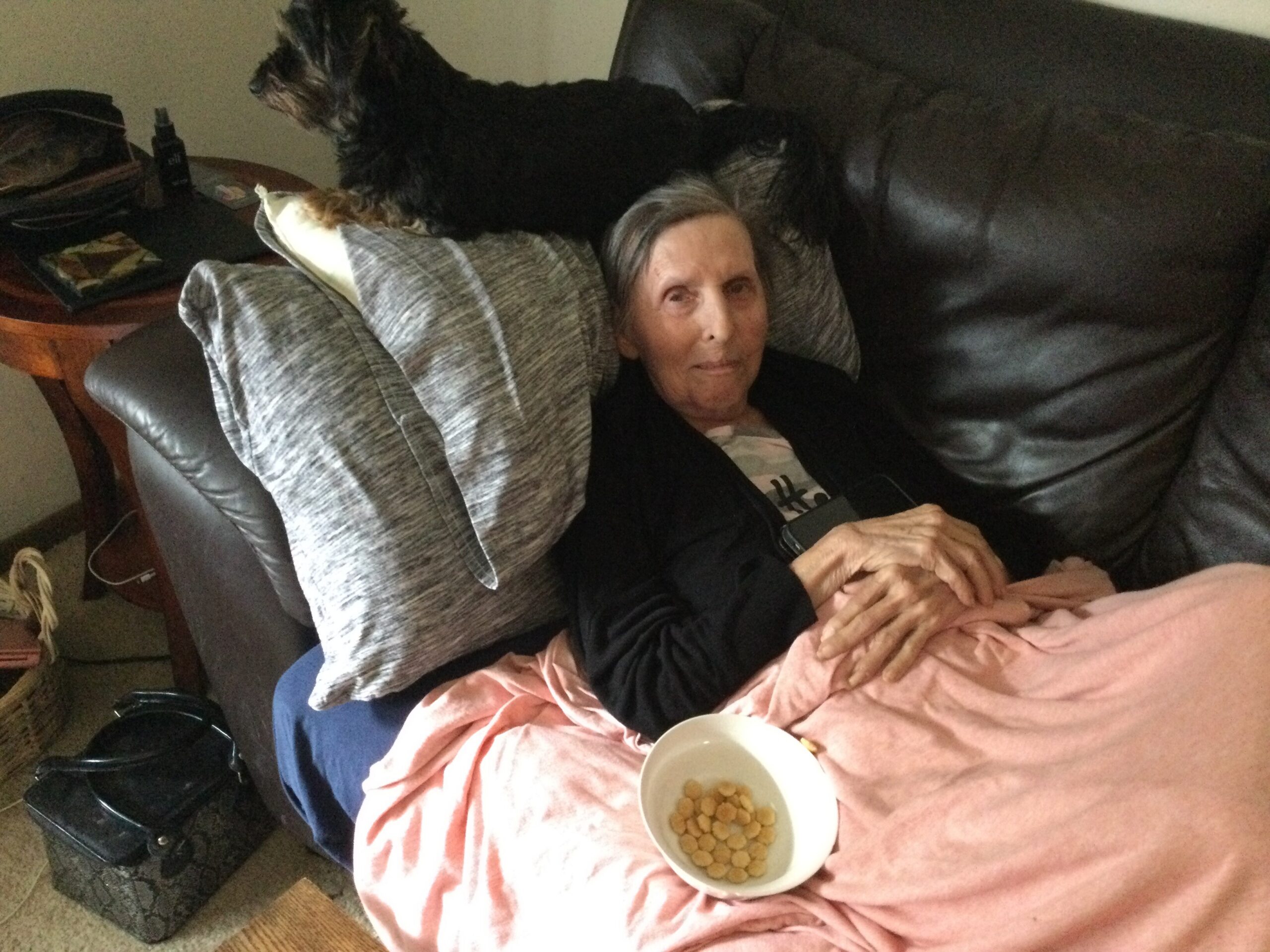 Elderly woman lying on a dark leather couch with a small black dog resting on her back; a bowl of round cereal sits on a pink blanket nearby.