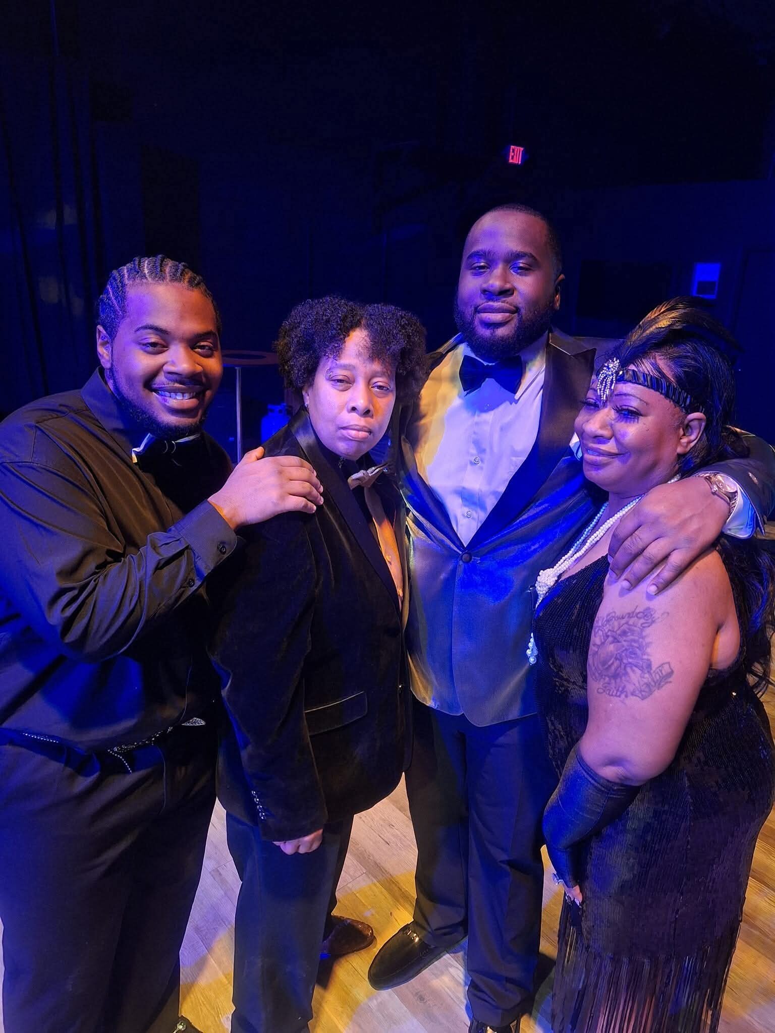 Four adults in formal wear pose together on a dimly lit stage with blue lighting, smiling at the camera while embracing each other.