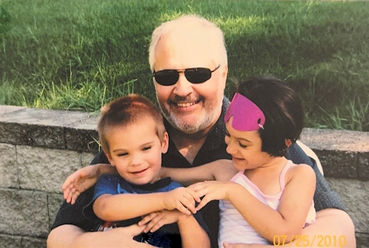 Smiling older man with sunglasses hugs a young boy and girl outdoors; girl wears a pink swim mask.
