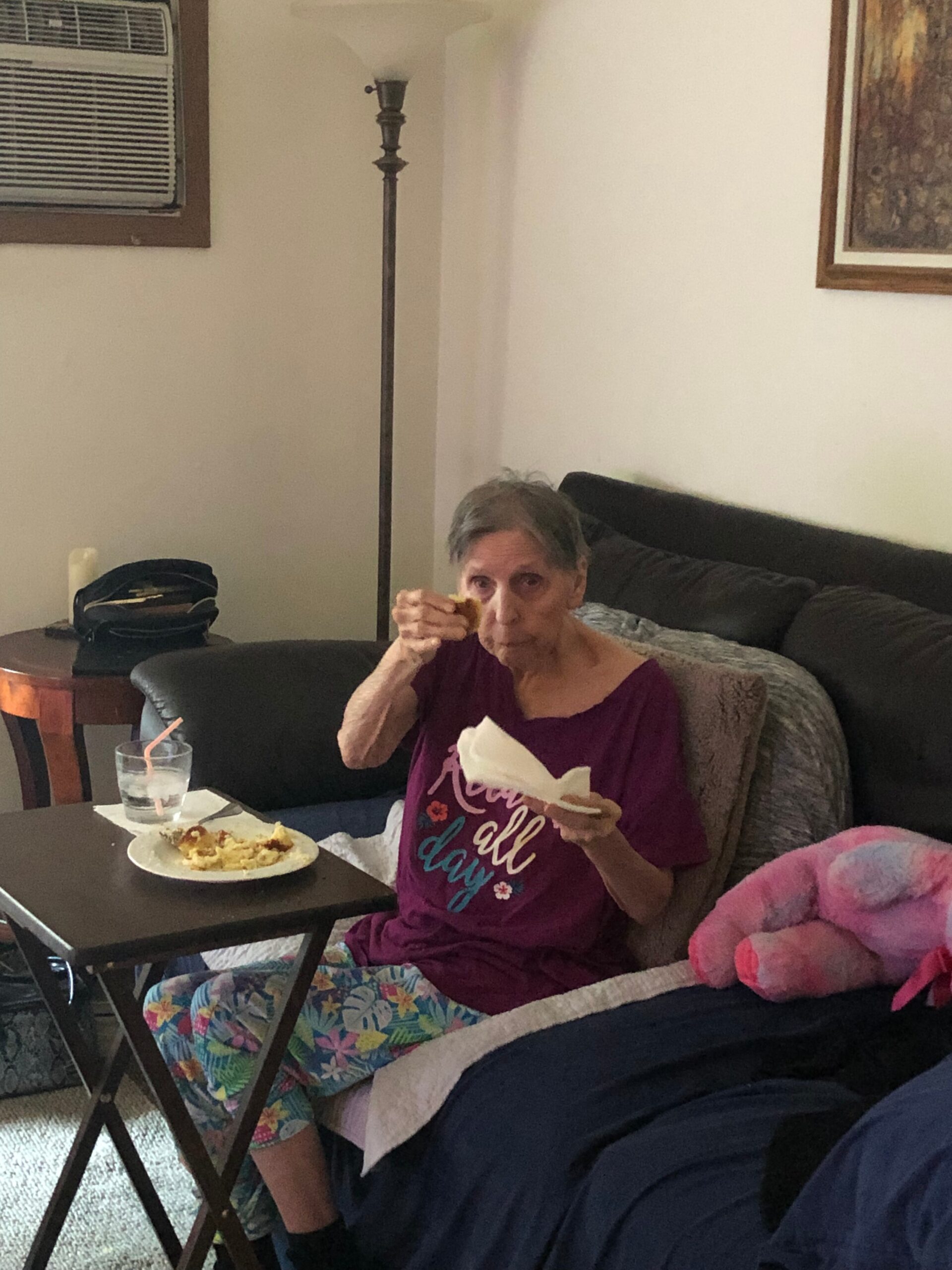 Older woman in a maroon shirt sitting on a couch, eating a slice of cake at a small table.