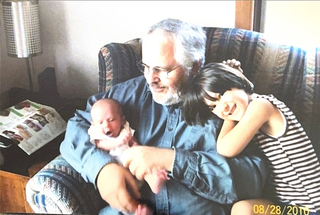 Older man with glasses cradling a newborn baby on his lap as a smiling young woman leans beside them on an armchair in a cozy living room.