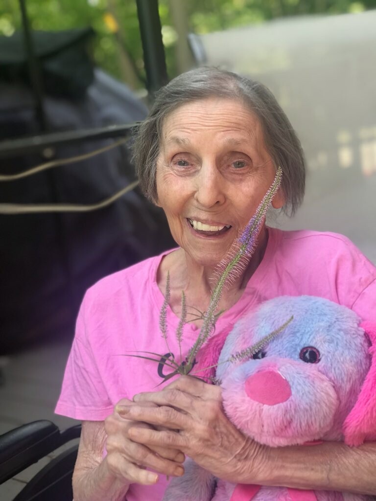 Smiling elderly woman in a pink shirt holds a purple plush bunny and a sprig of purple flowers outdoors.