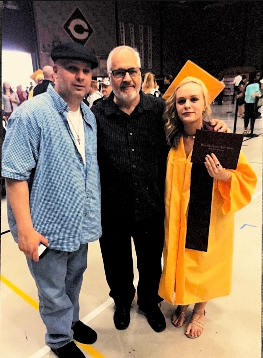 Graduate in orange cap and gown posing with two men at a graduation ceremony, holding a diploma.