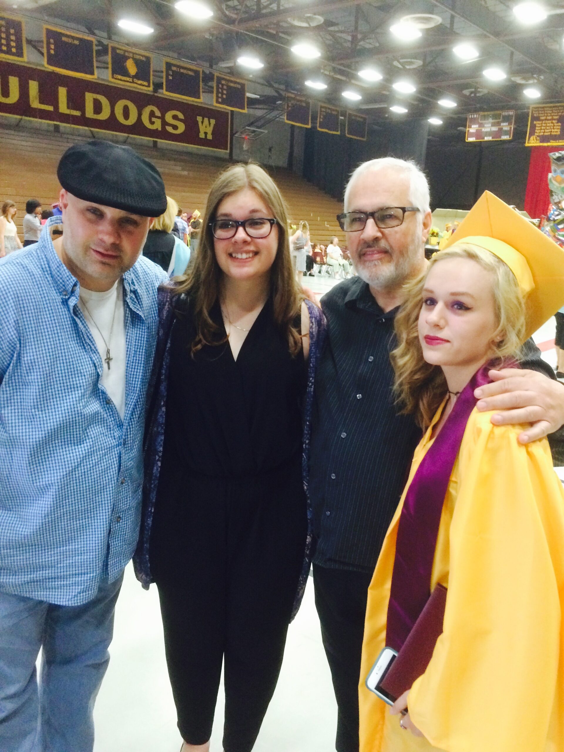 Four people posing for a graduation photo in a gymnasium; the graduate wears a yellow cap and gown with a maroon stole, flanked by three family members in casual clothes.