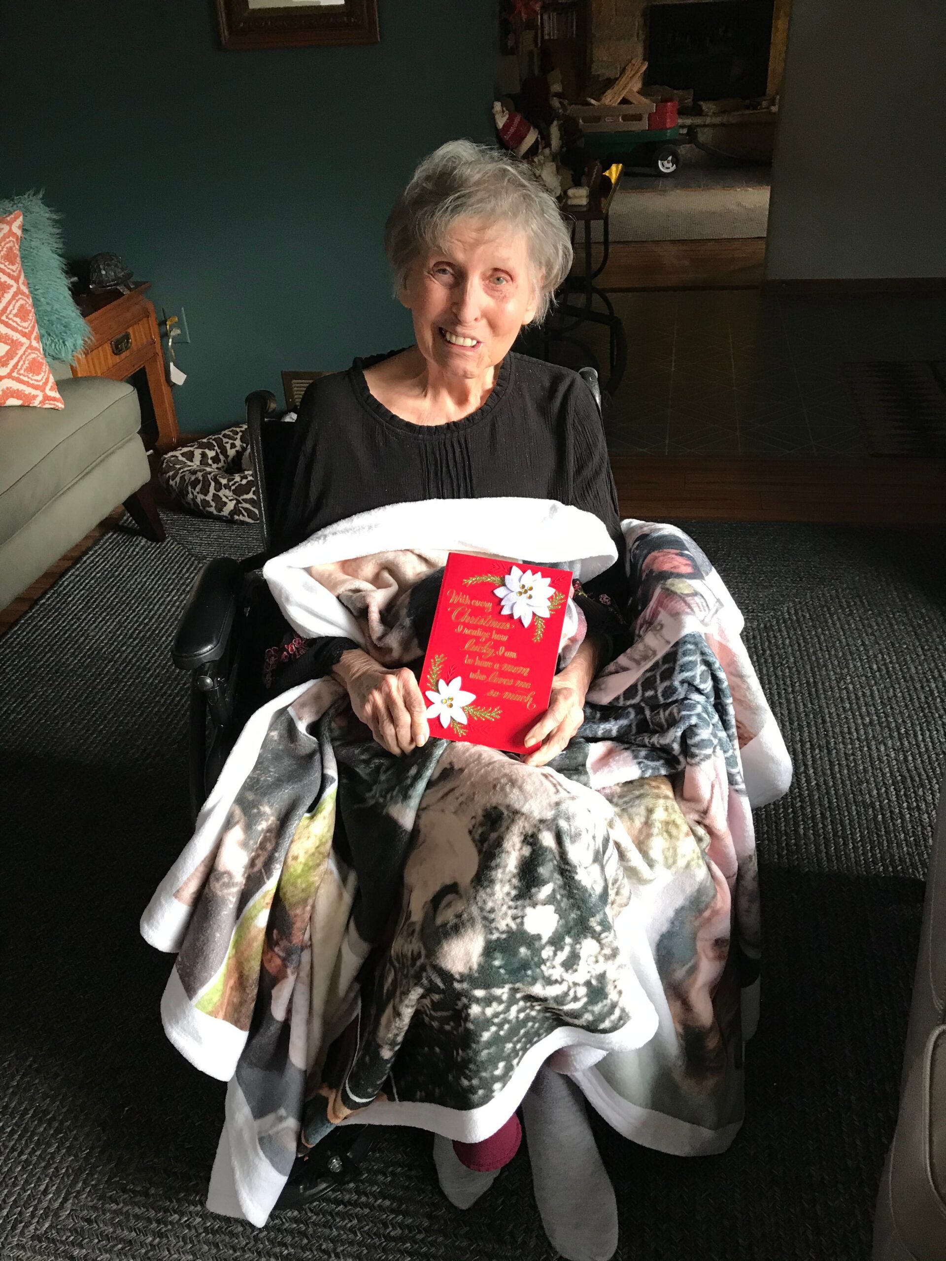 Smiling elderly woman in a wheelchair wrapped in a blanket, holding a red Christmas card indoors.