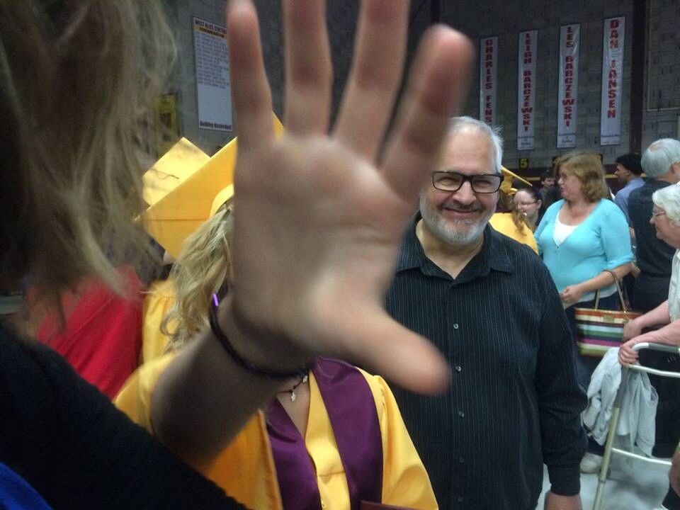 Smiling man with glasses in a dark shirt among a crowd; a raised hand fills the foreground, partially obscuring the camera.