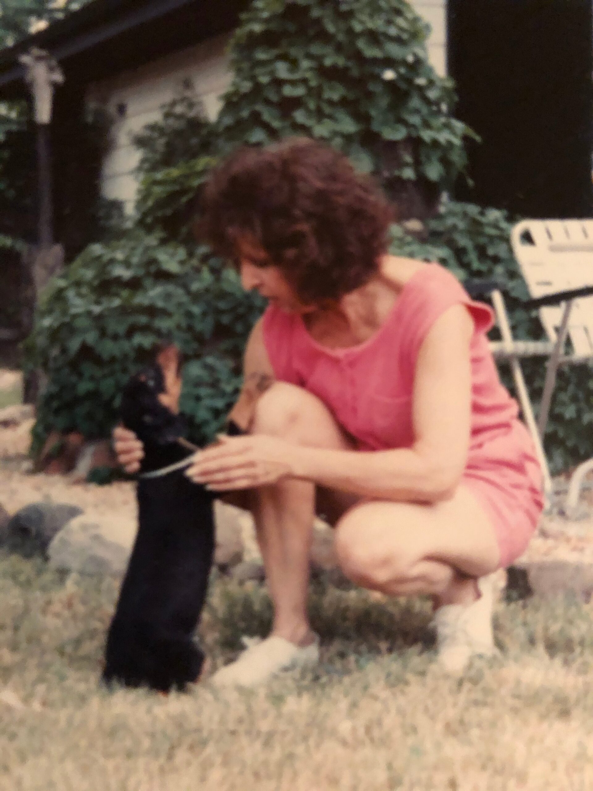 Person with curly brown hair wearing a pink sleeveless outfit crouches in a garden, looking down at something in their hands, ivy and a white chair nearby in the background.