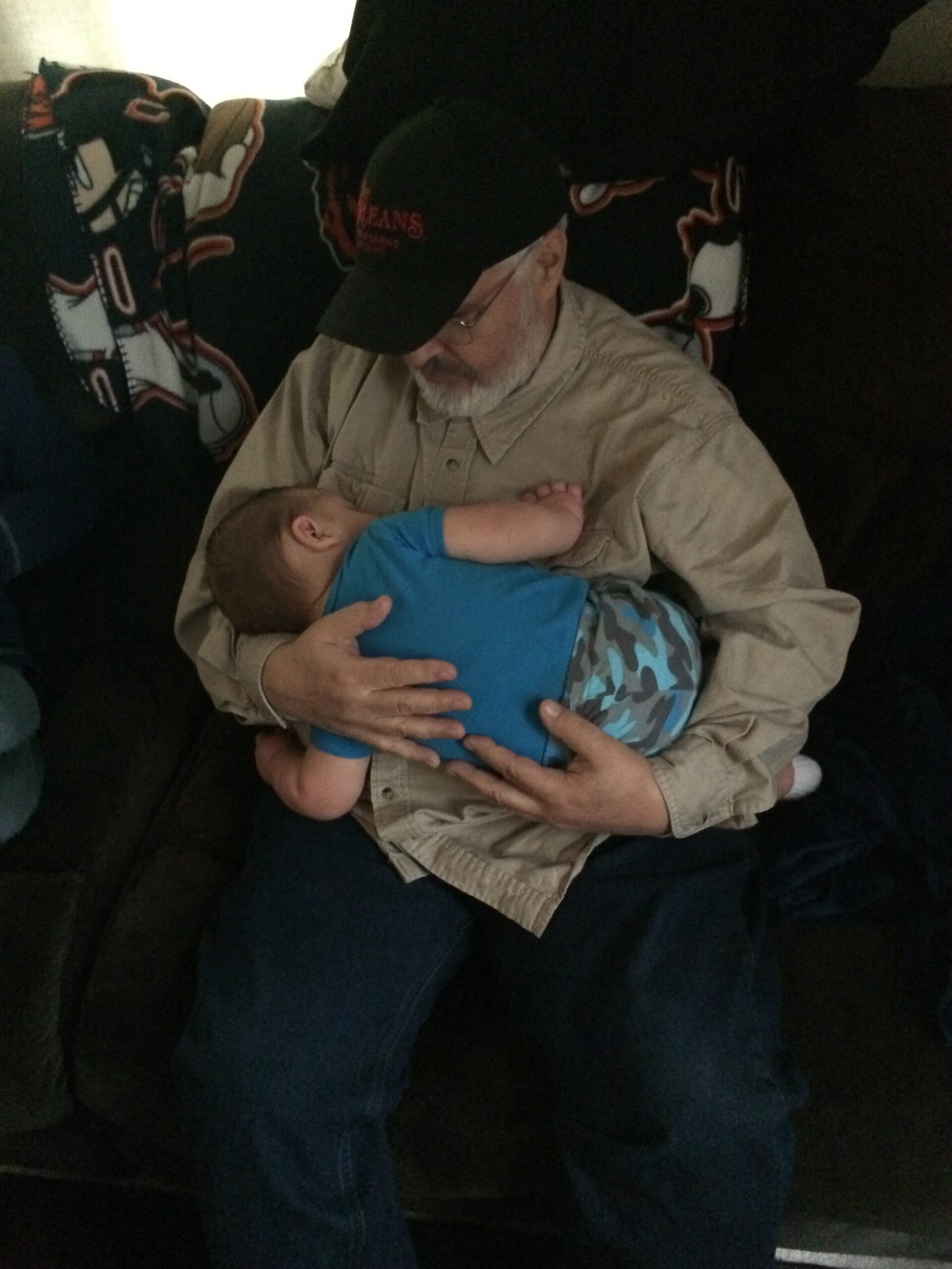 Older man with a cap and glasses cradling a baby on a dark couch, both resting against a patterned blanket background on the back of the sofa.