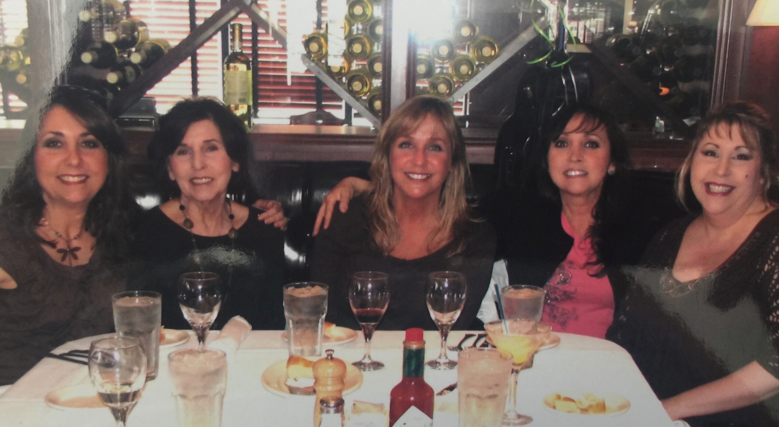 Five women sit together at a restaurant table, smiling for the camera with drinks and plates in front of them.