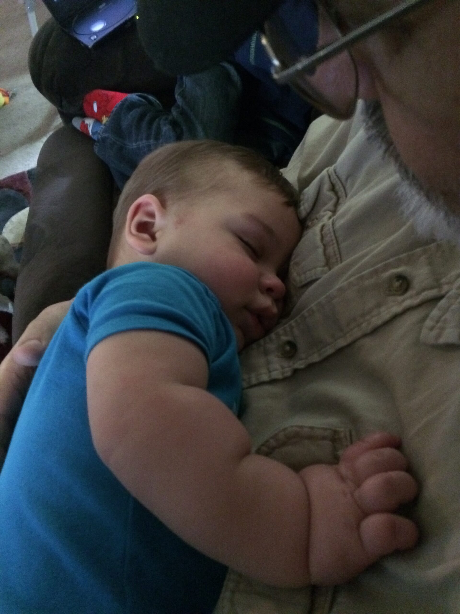 Sleeping infant in a blue shirt cradled on a caregiver's chest.