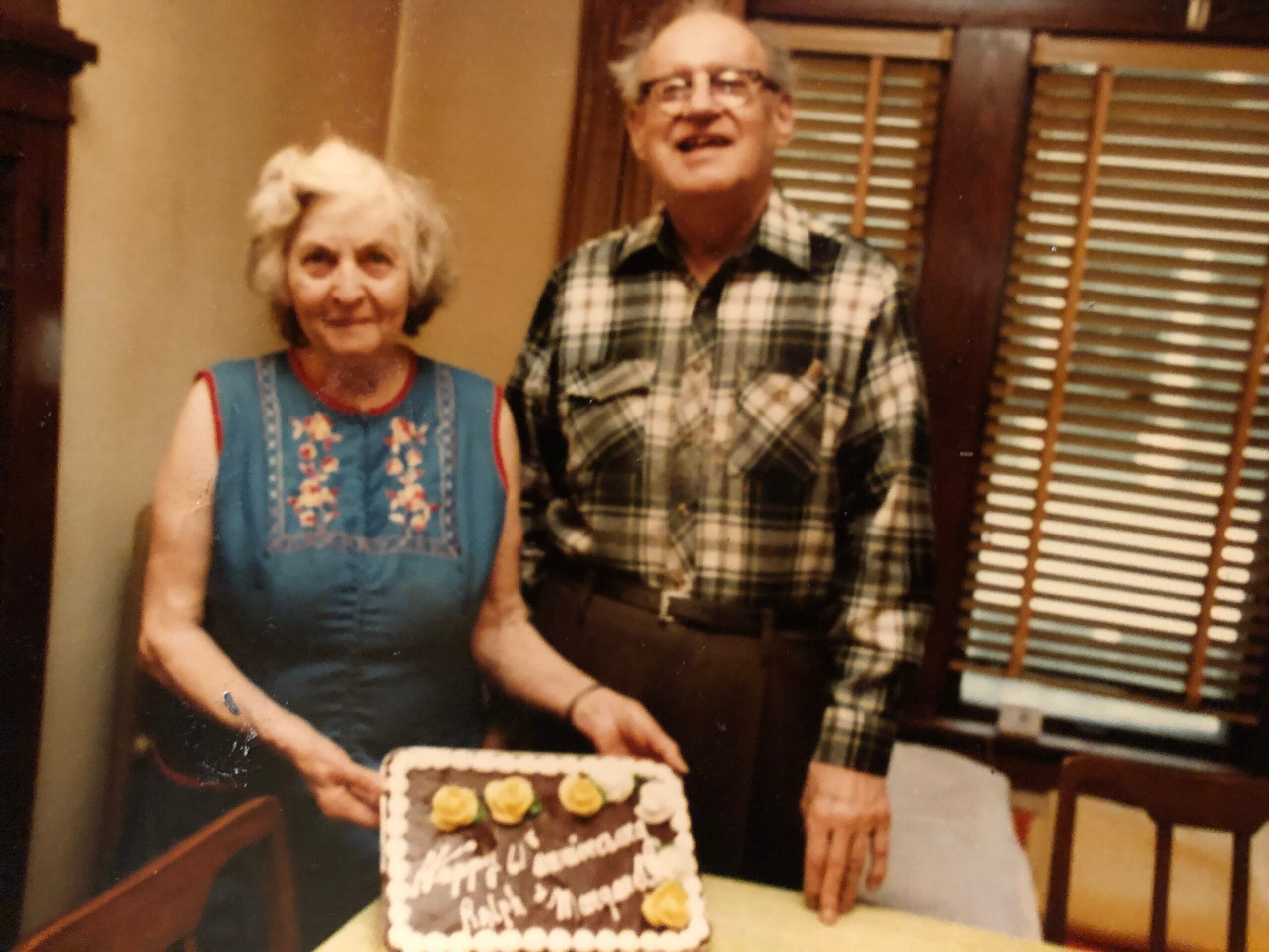 Two elderly adults standing side by side with a frosted birthday cake in front of them, smiling in a home setting.