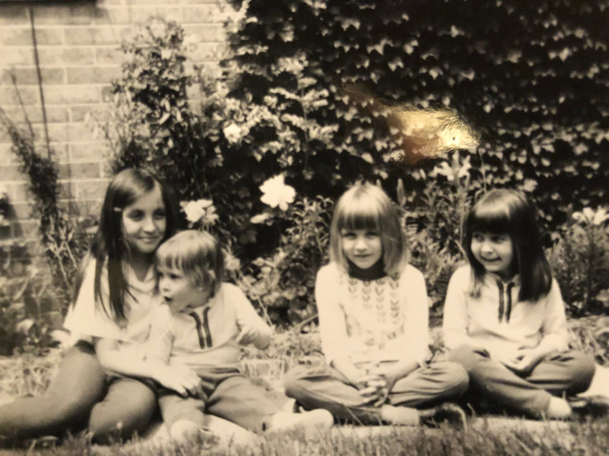 Black-and-white photo of five young girls sitting on the grass in a garden, with ivy and a brick wall behind them, looking at the camera.