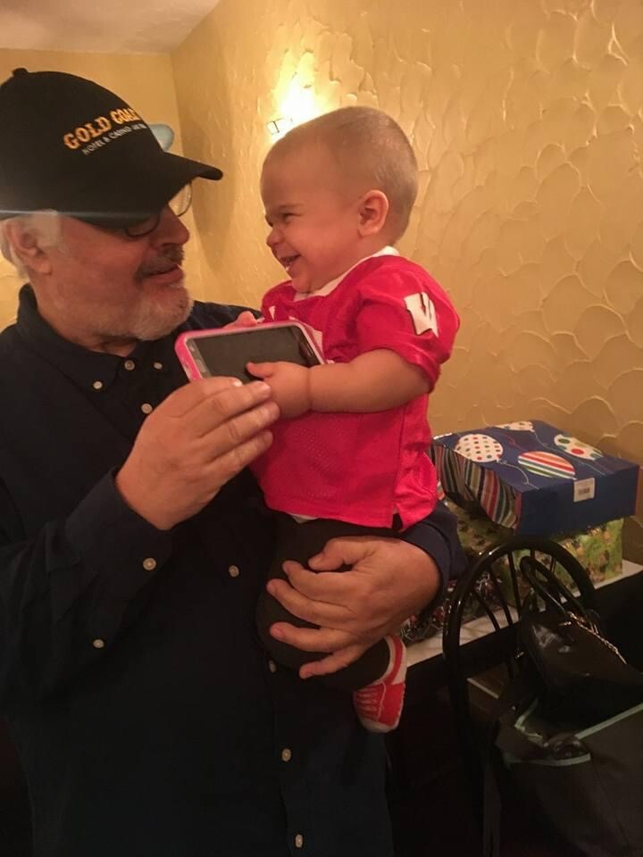 Older man in a cap holds a smiling baby wearing a red shirt indoors, near a wall with textured wallpaper.