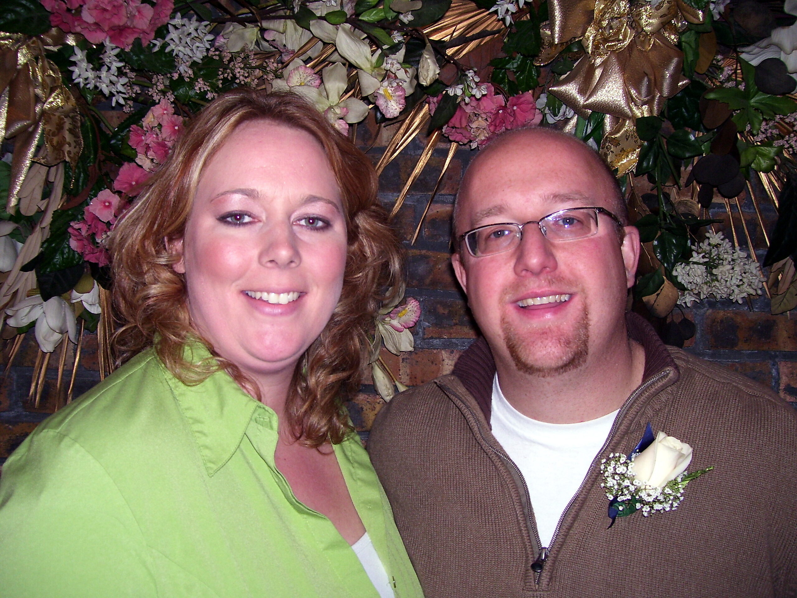 Smiling couple posing for a selfie in front of a floral decorative backdrop with pink flowers and gold ribbons.