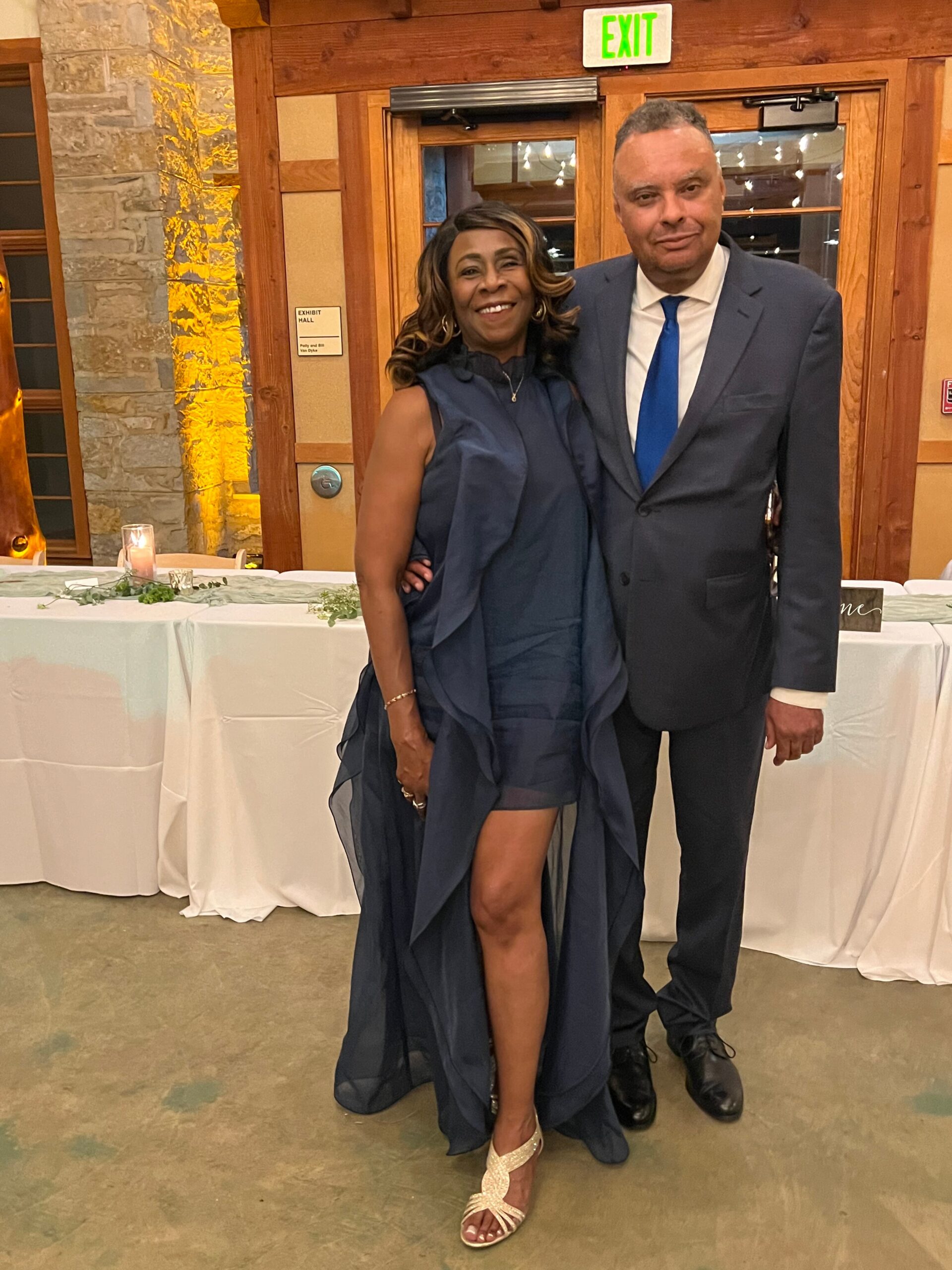 Smiling couple posing at a formal event; woman in a navy gown with a high slit and the man in a dark suit with a blue tie, banquet tables in the background.