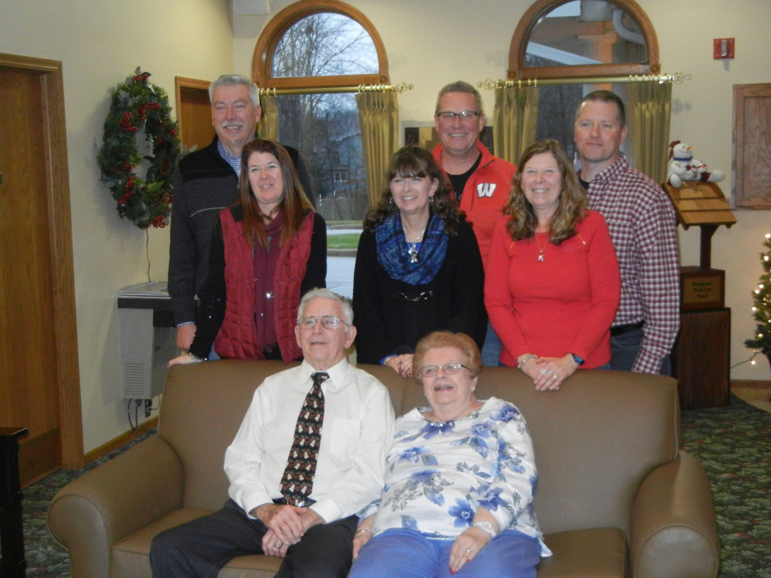Group of eight adults in a holiday-decorated living room, two seniors seated on a brown sofa while six others stand behind them.