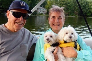 Two people on a boat smiling at the camera, holding two small white dogs in yellow life jackets on a lake.