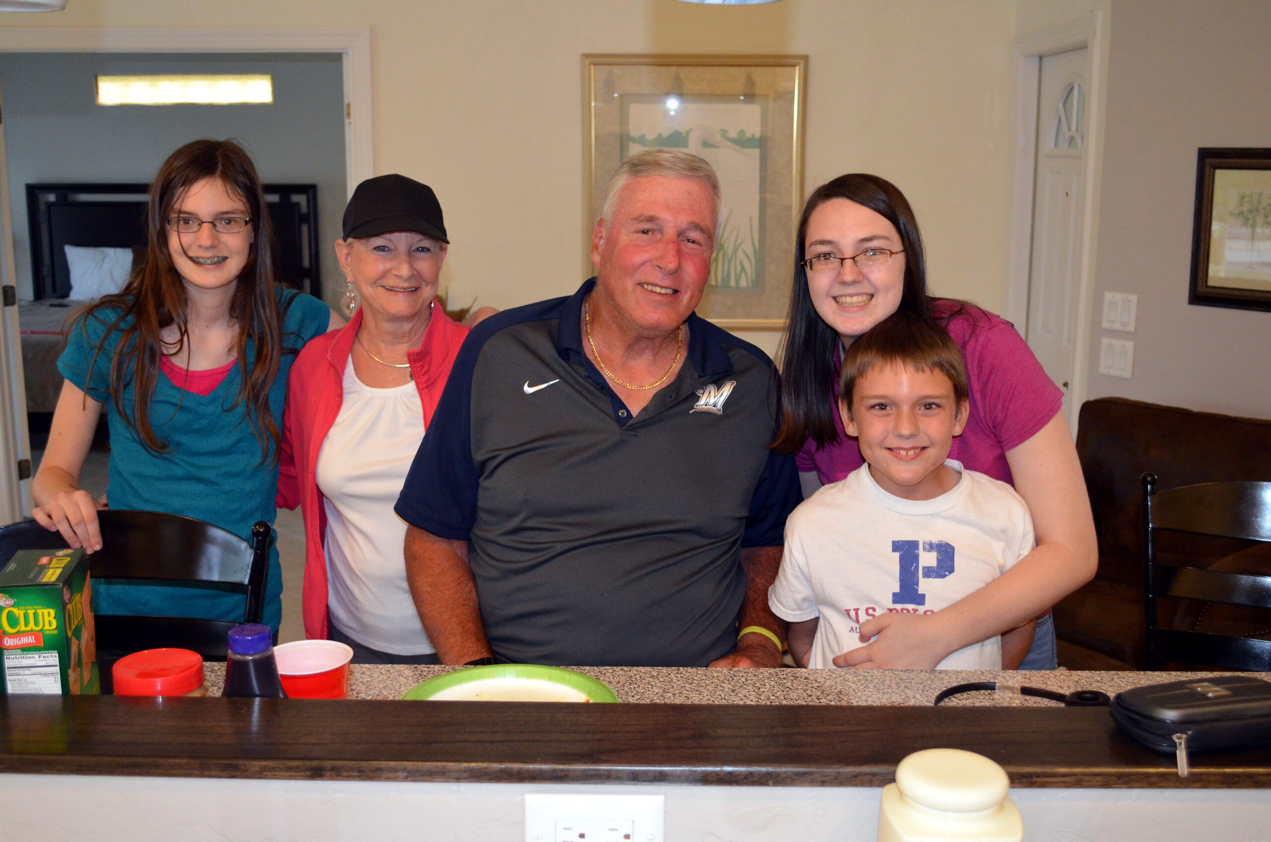 Five people smiling at a kitchen counter: a teenage girl with braces, an older woman, a man, a woman in pink, and a boy.