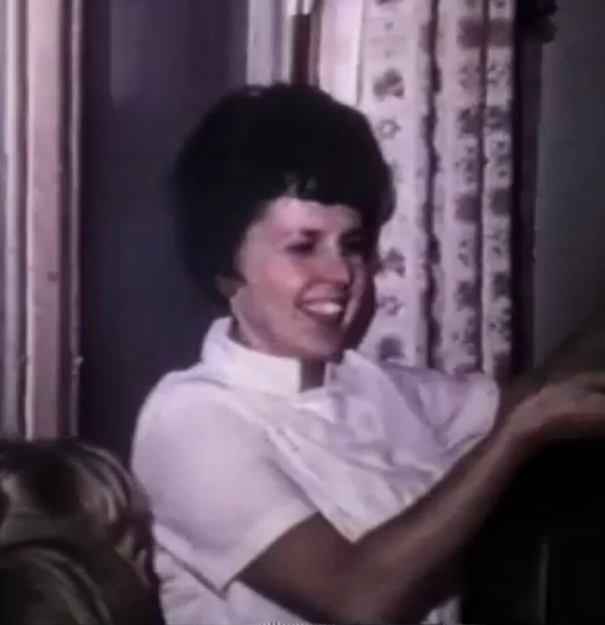 Smiling woman with dark hair in a white shirt, indoors near patterned curtains, mid-conversation or laughter scene.