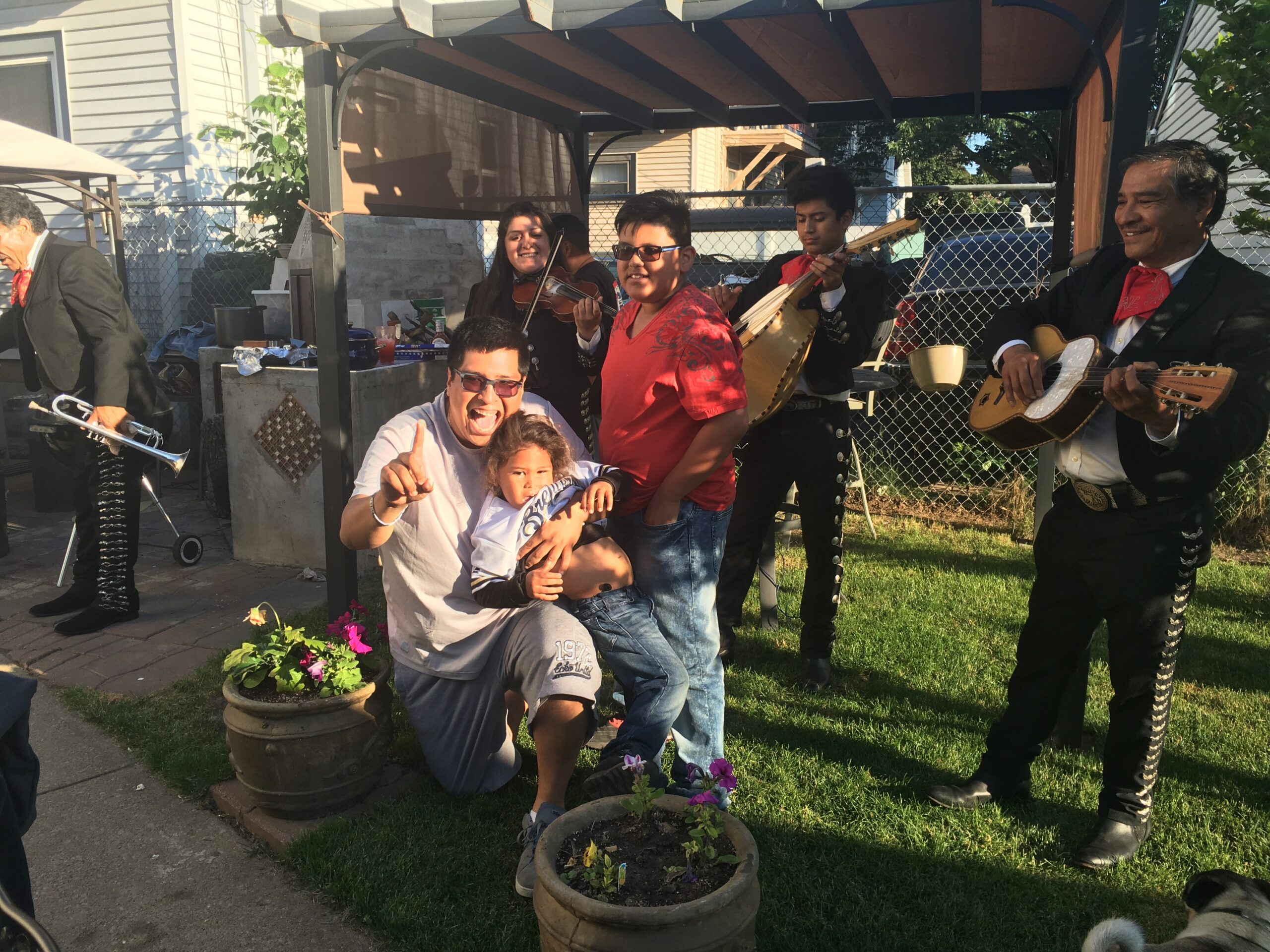 Family and mariachi band posing outdoors under a canopy; two kids up front, adults beside them, musicians play guitar and violin in a backyard setting.