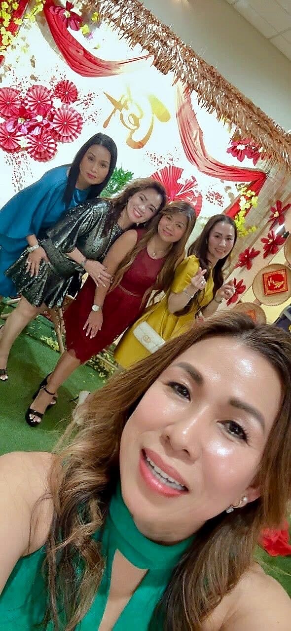 Group of five women posing for a selfie at a festive event with red and gold decorations in the background.