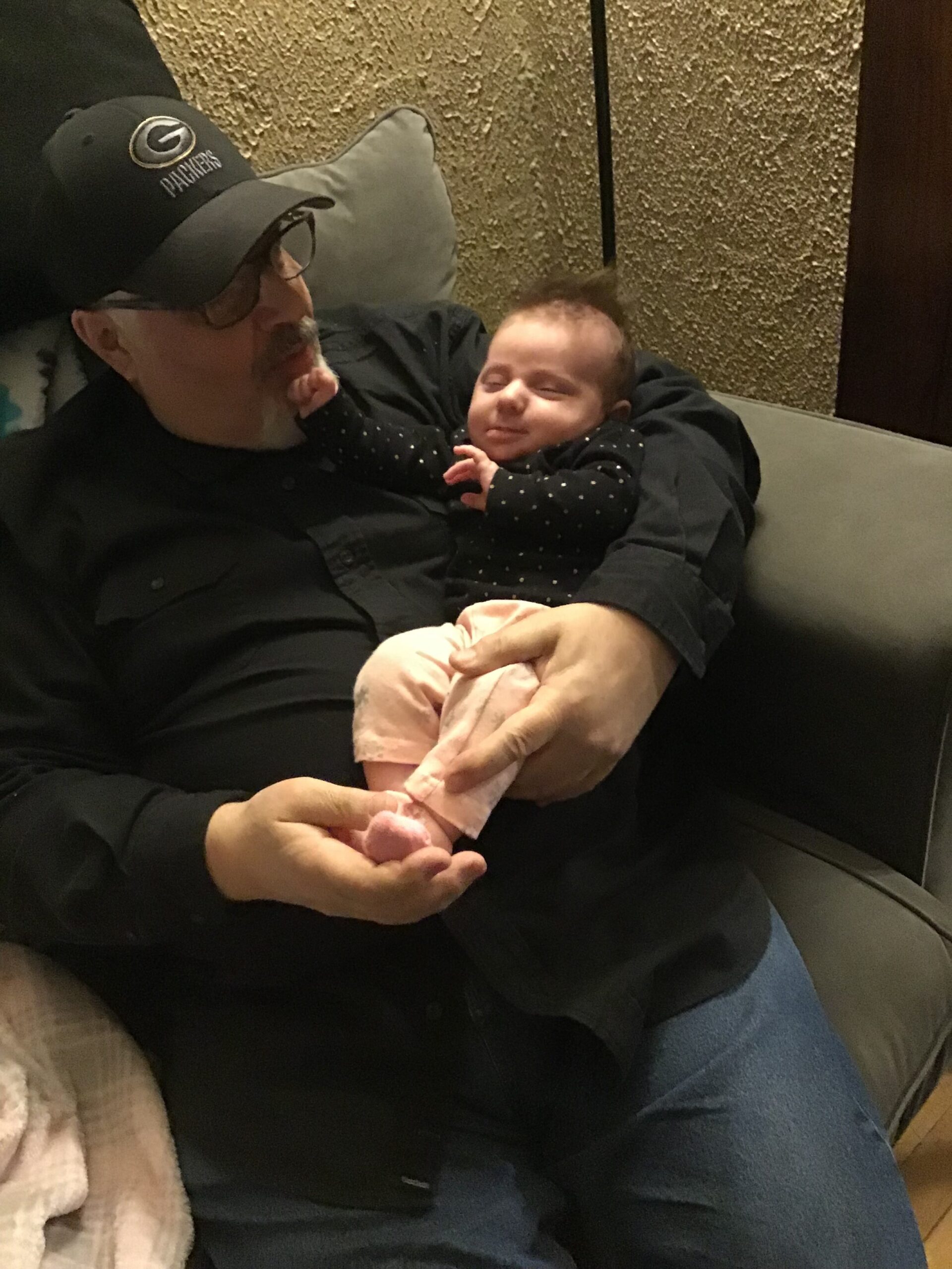 An older man with glasses and a Packers cap sits on a couch, cradling a smiling baby in his arms.