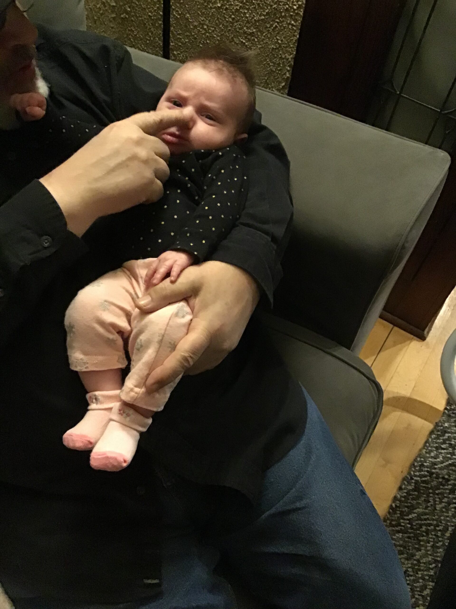 Adult gently cradling a sleeping baby on a dark sofa; baby wears a navy polka-dot onesie and pink pants with pink socks.