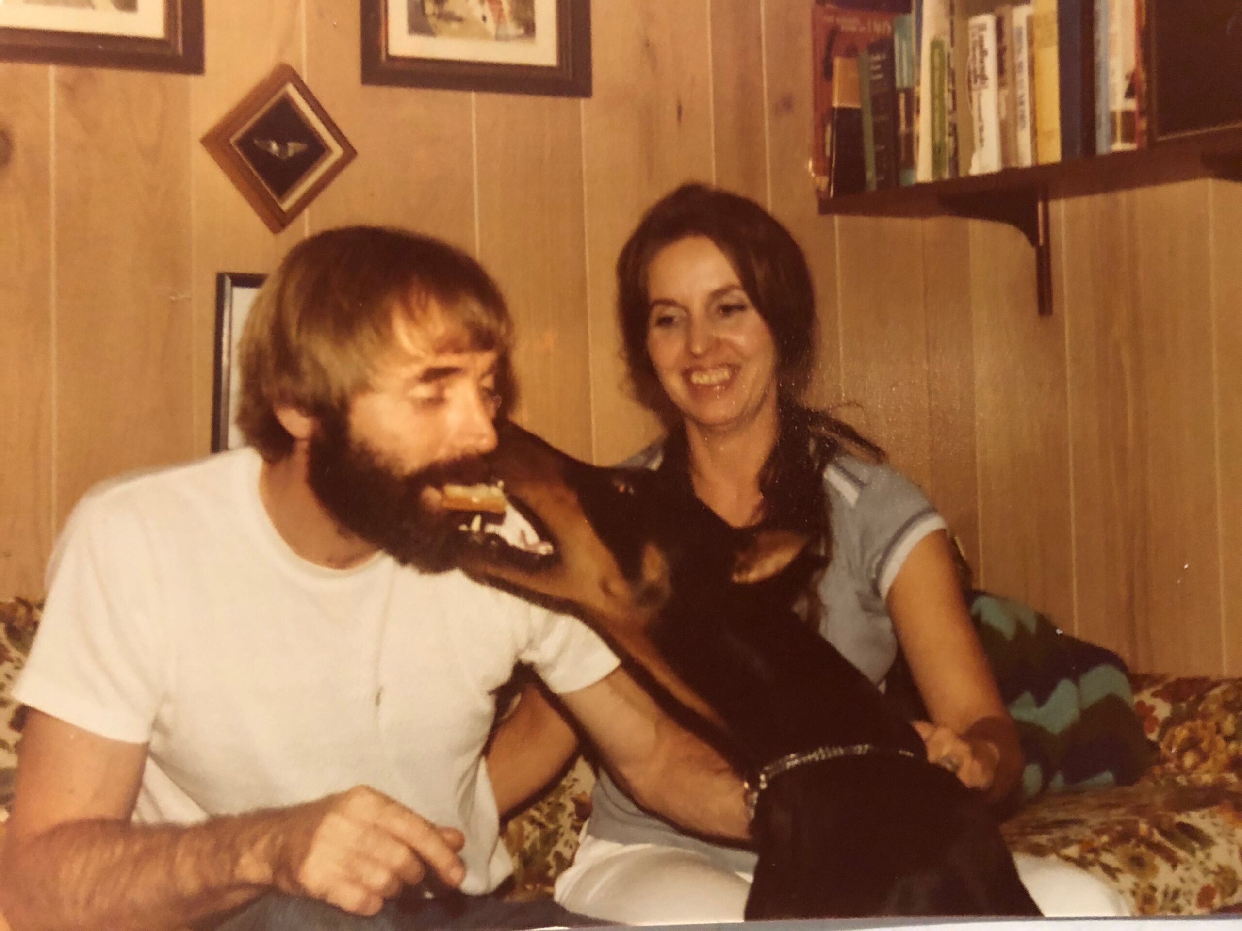 Two friends sit closely together indoors, smiling at the camera; wood-paneled walls and a bookshelf visible behind them.