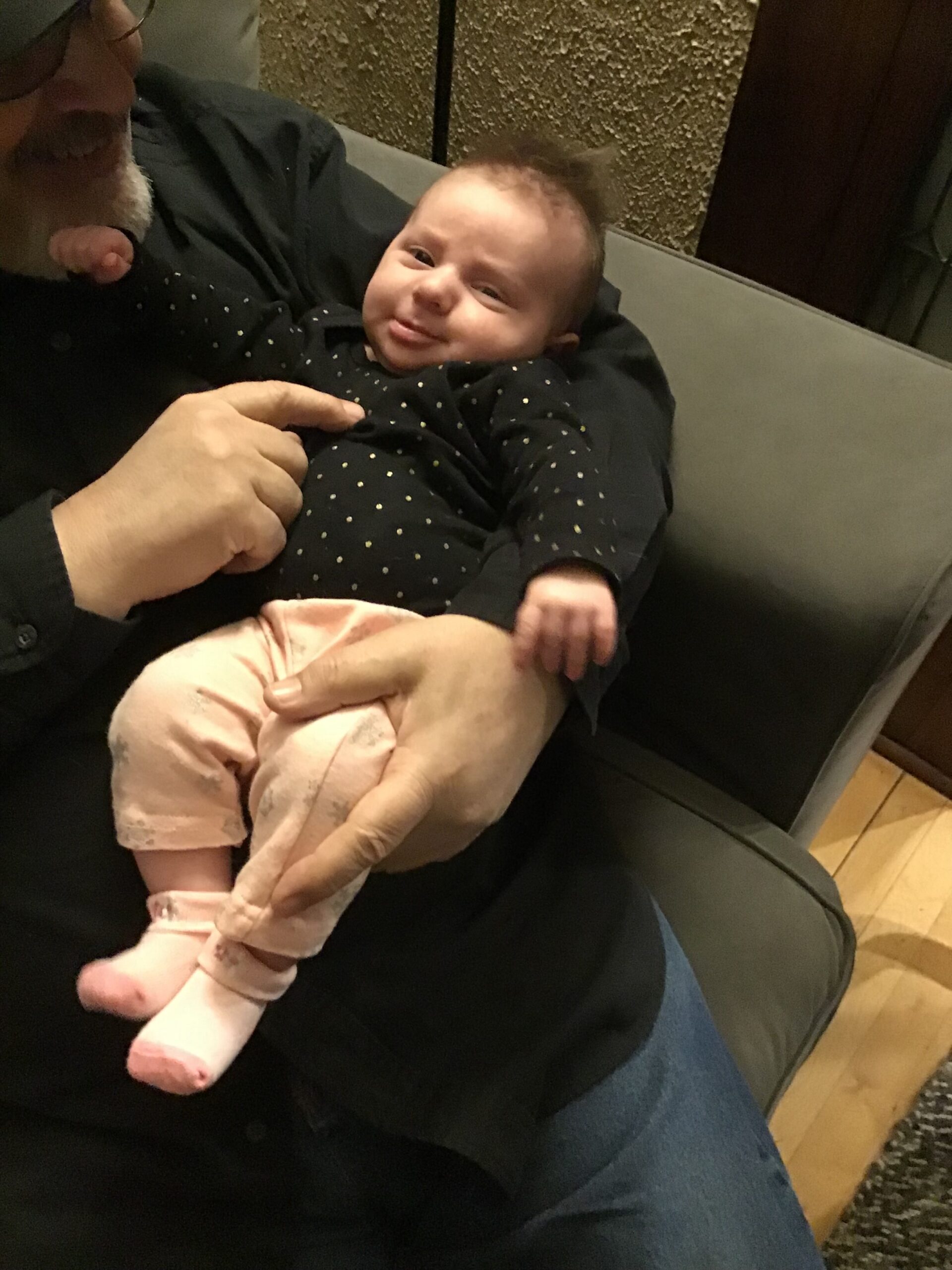 Smiling baby in a dark polka-dot onesie being held on a black couch by an adult's hands and arm visible in the frame.