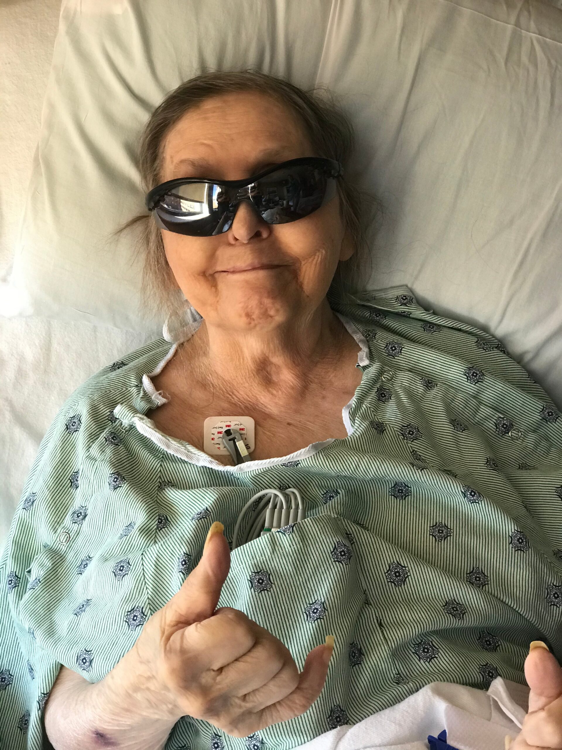 Older woman in a hospital bed wearing dark protective glasses and a patterned gown, with a chest monitor, giving a thumbs-up gesture.
