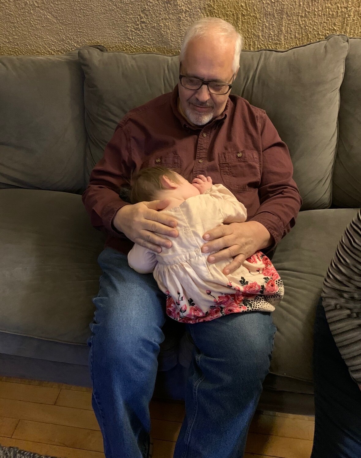 Older man with white hair and glasses sits on a gray couch, cradling a small child in a pink floral dress.