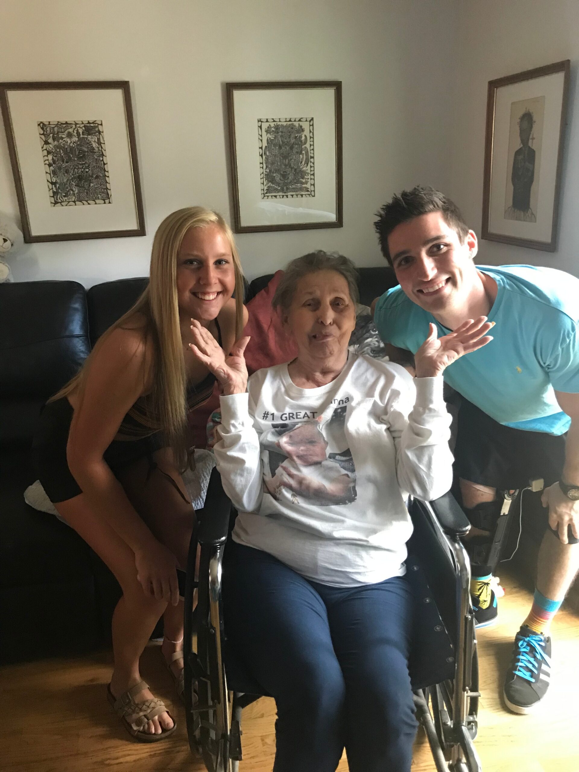 Smiling woman kneels beside an elderly woman in a wheelchair, with a smiling man leaning in from the right in a living room.