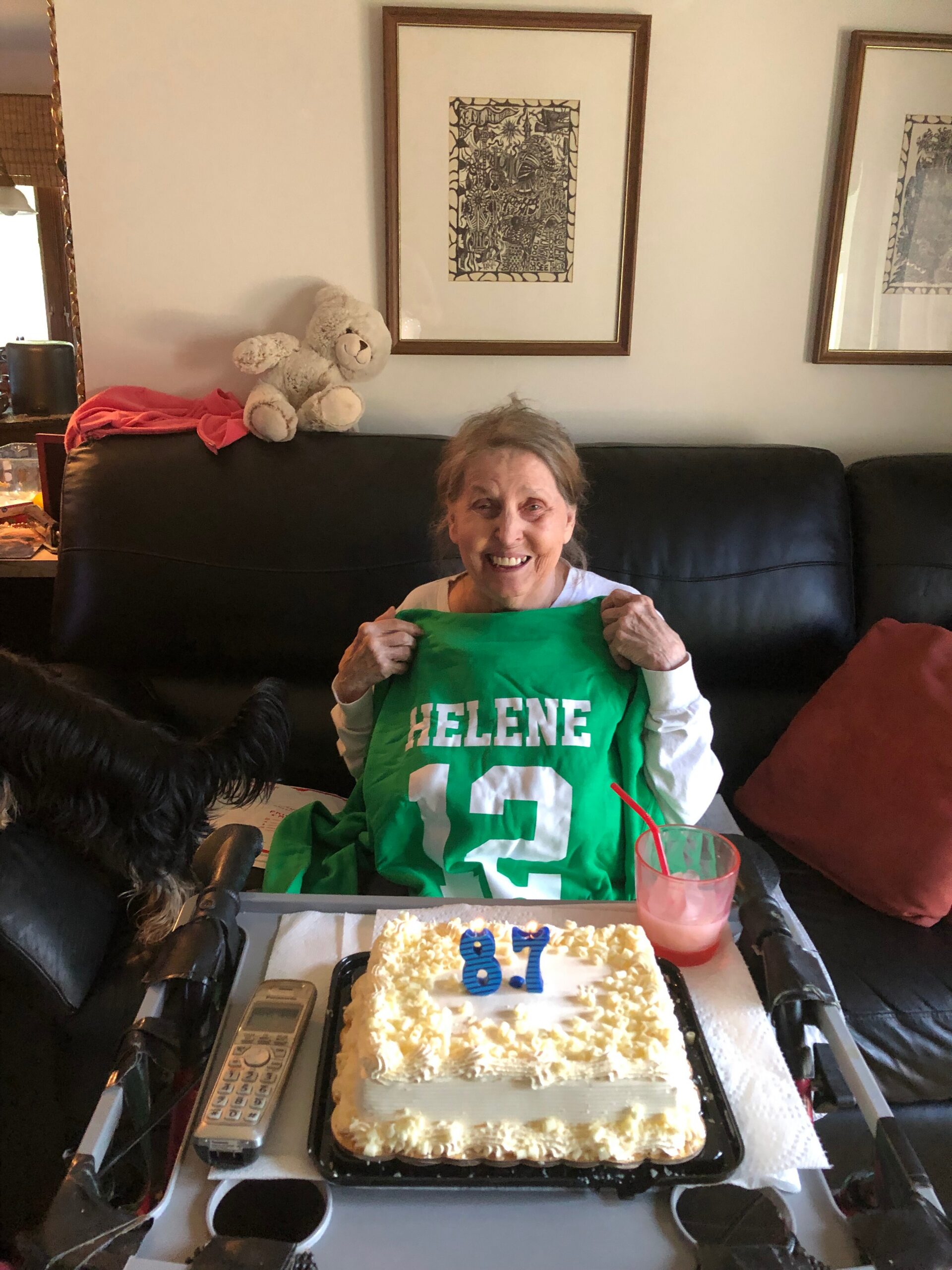 Smiling elderly woman sitting on a black sofa, holding a green birthday shirt that reads 'HELENE 13'; a cake with blue '87' candles sits in front, with a red drink and remote on the table nearby.
