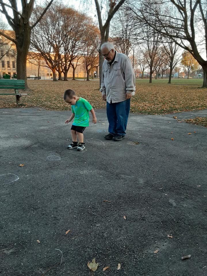 Grandfather watches grandson draw chalk marks on the park sidewalk in autumn light.