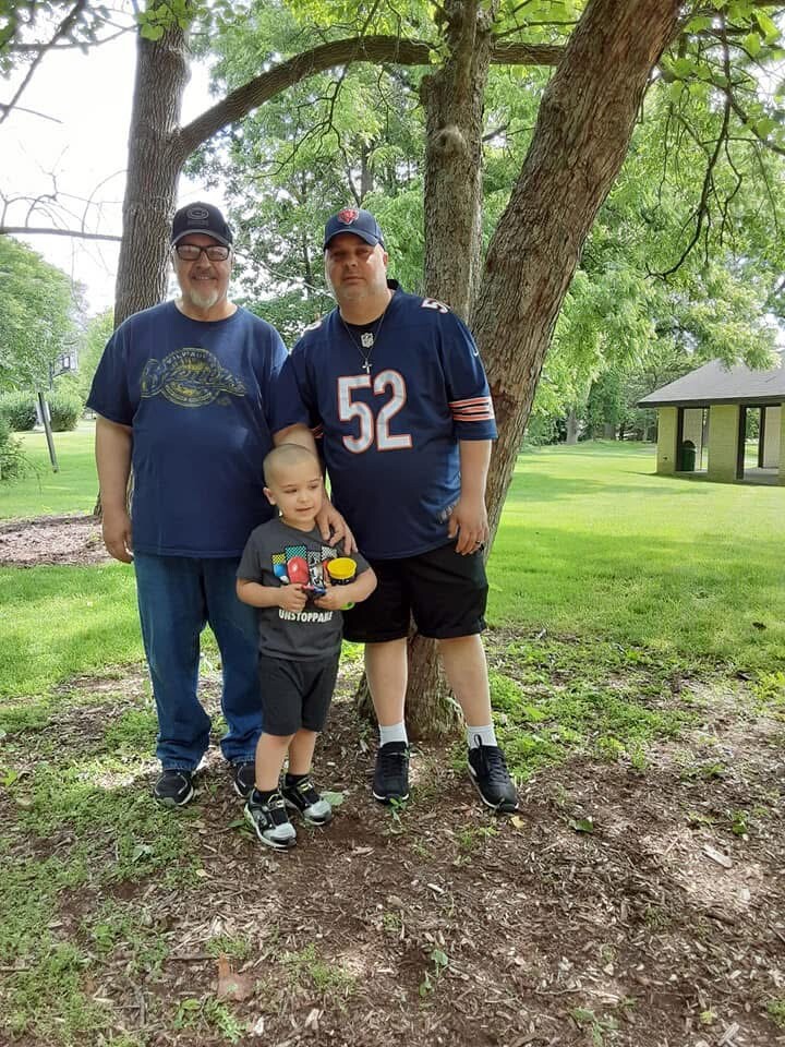 Three people posing under a tree in a park: an older man in a blue shirt, a man in a Chicago Bears jersey (52), and a small boy holding toys with the tree behind them.