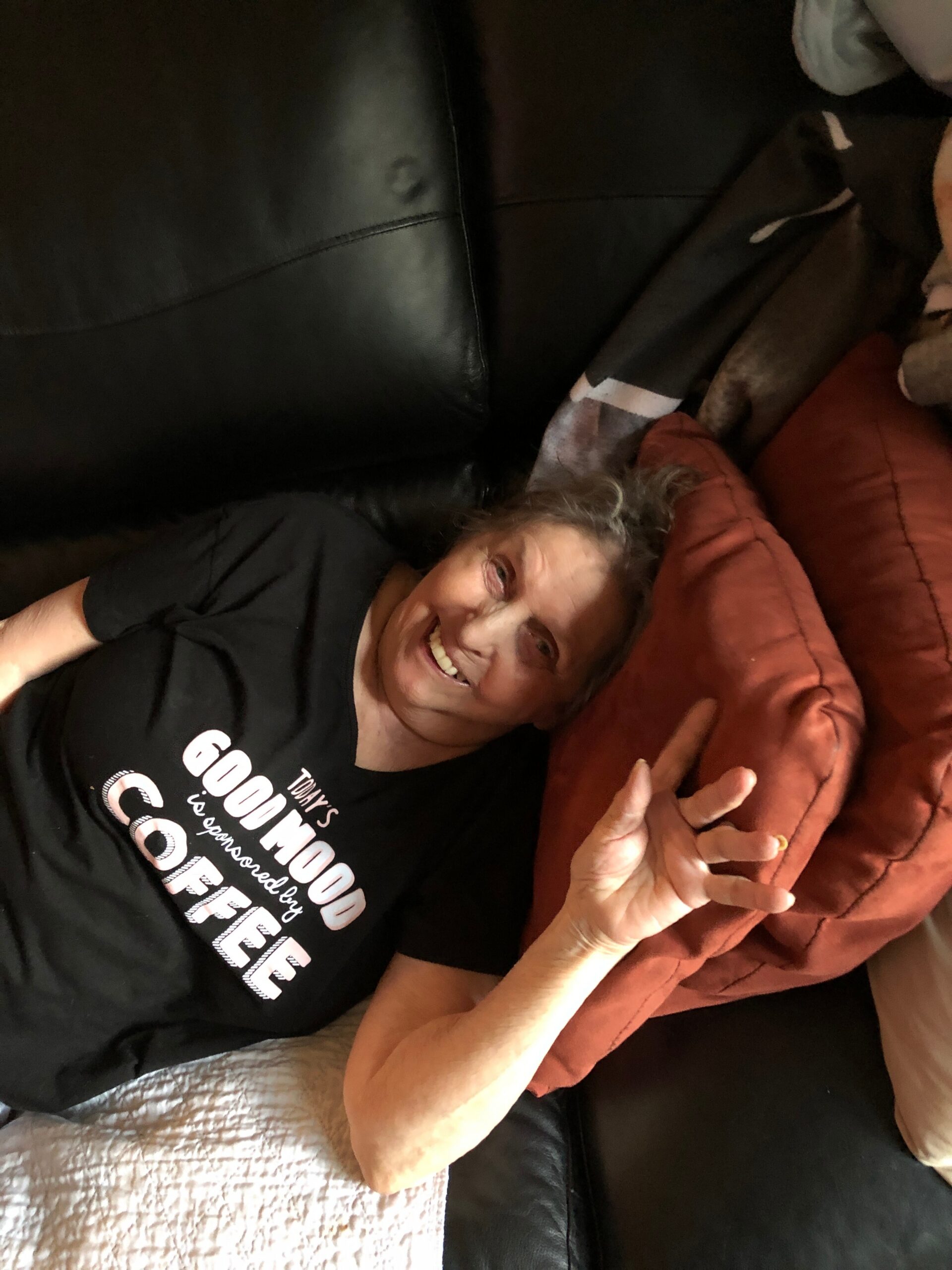 Smiling older person reclining on a black leather couch, with a brown blanket beside them.