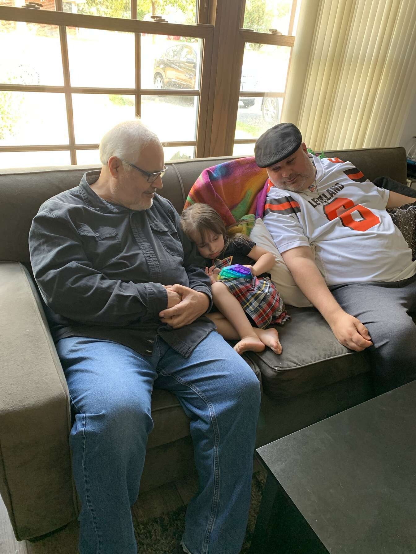 Older man with glasses sits on left side of sofa, smiling at a young girl cuddled between him and a man in a cap; they rest together indoors near a window.
