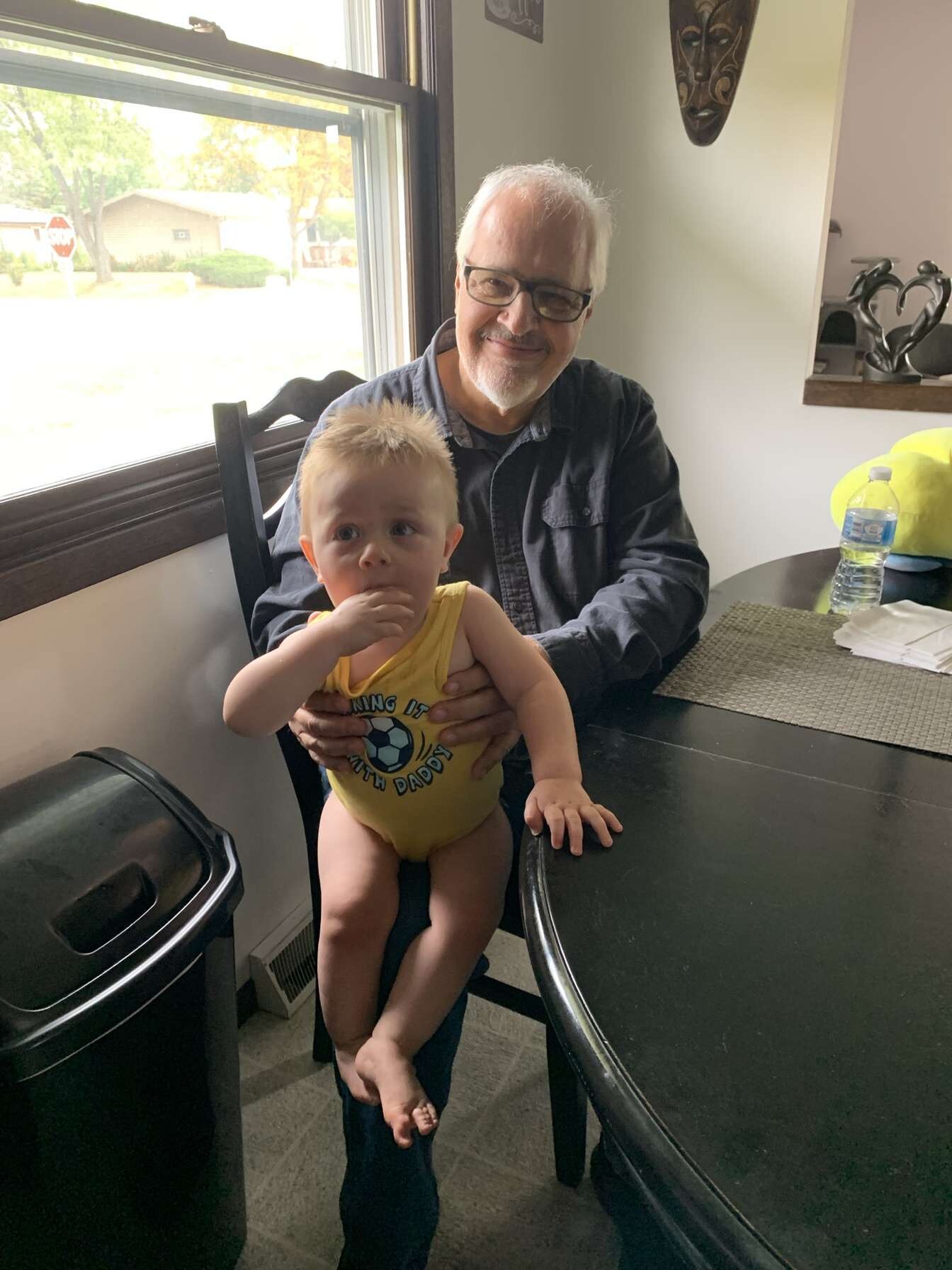 Smiling older man with glasses holds a baby on a chair at a dining table by a window in a home.