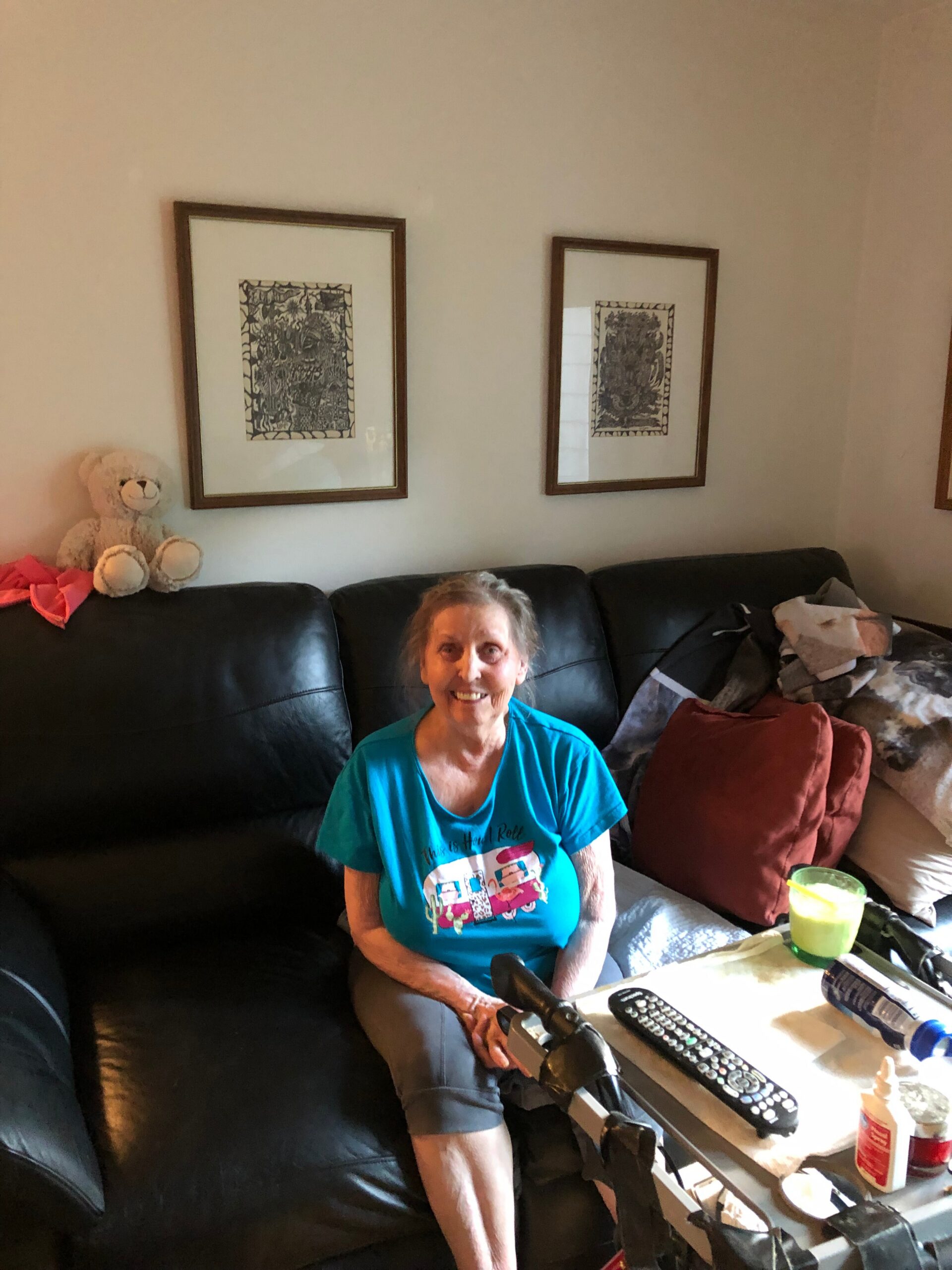 Smiling elderly woman in a turquoise shirt sits on a black leather sofa in a living room with two framed artworks on the wall and a teddy bear nearby.