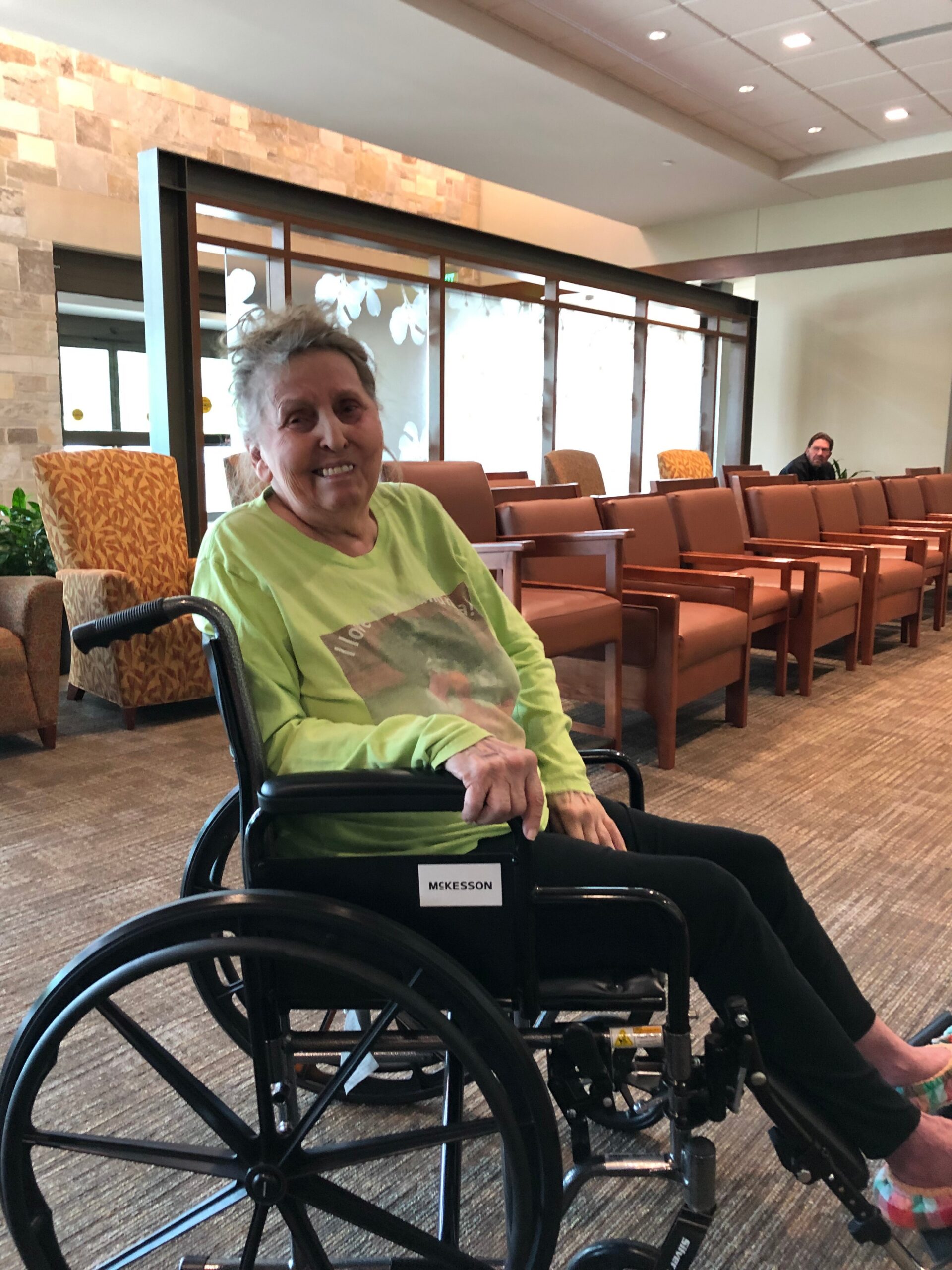 Smiling elderly woman in a green shirt sits in a wheelchair in a waiting area with brown chairs behind her, MSKesson on the chair label.
