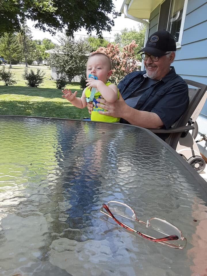 Grandfather holds a baby on a sunny porch, baby with pacifier in a bright green onesie; sunglasses lay on a glass outdoor table, suburban yard in background.