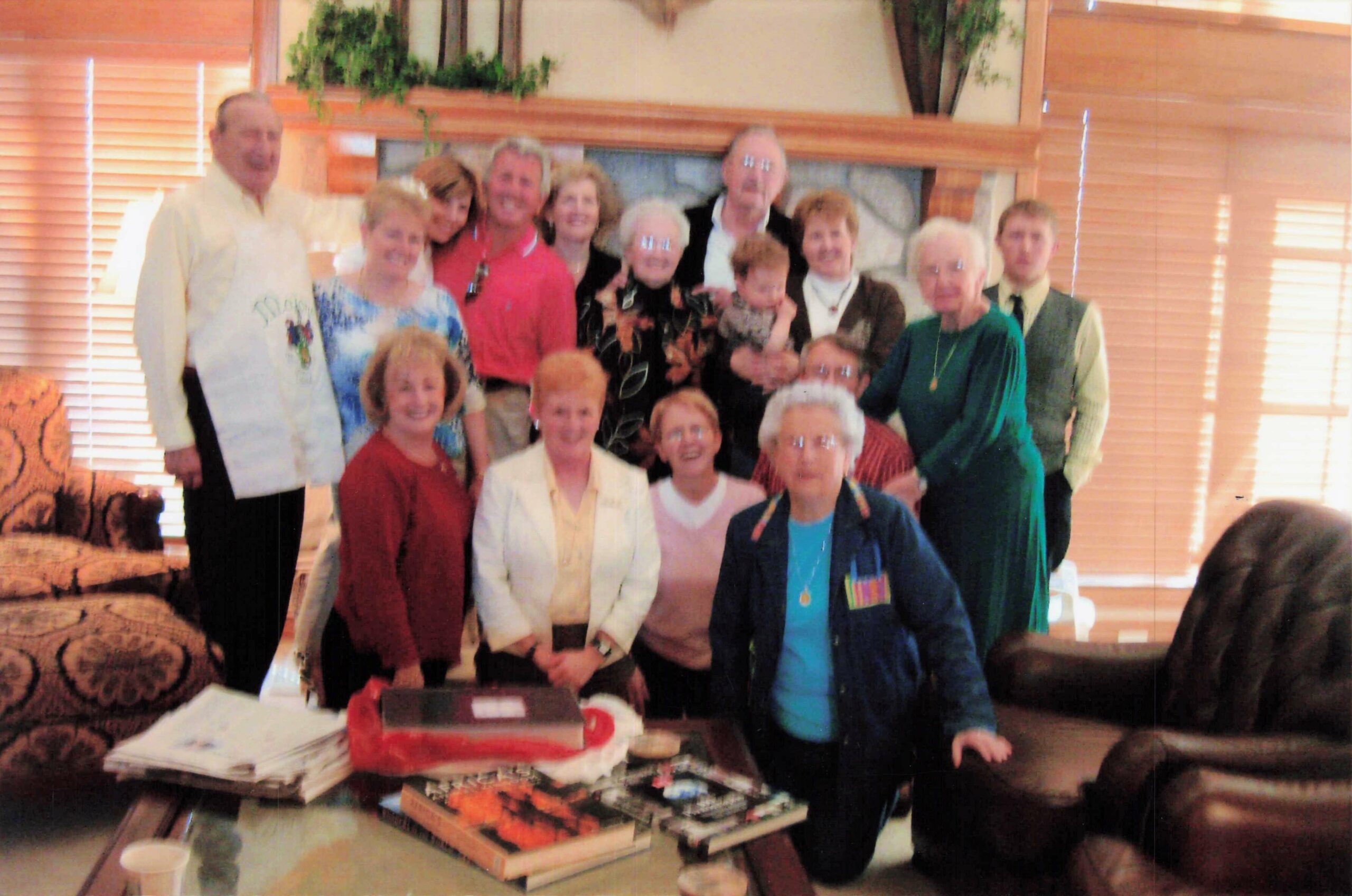 A group of about 15 adults posing in a living room around a table with a birthday cake, smiling for the photo.