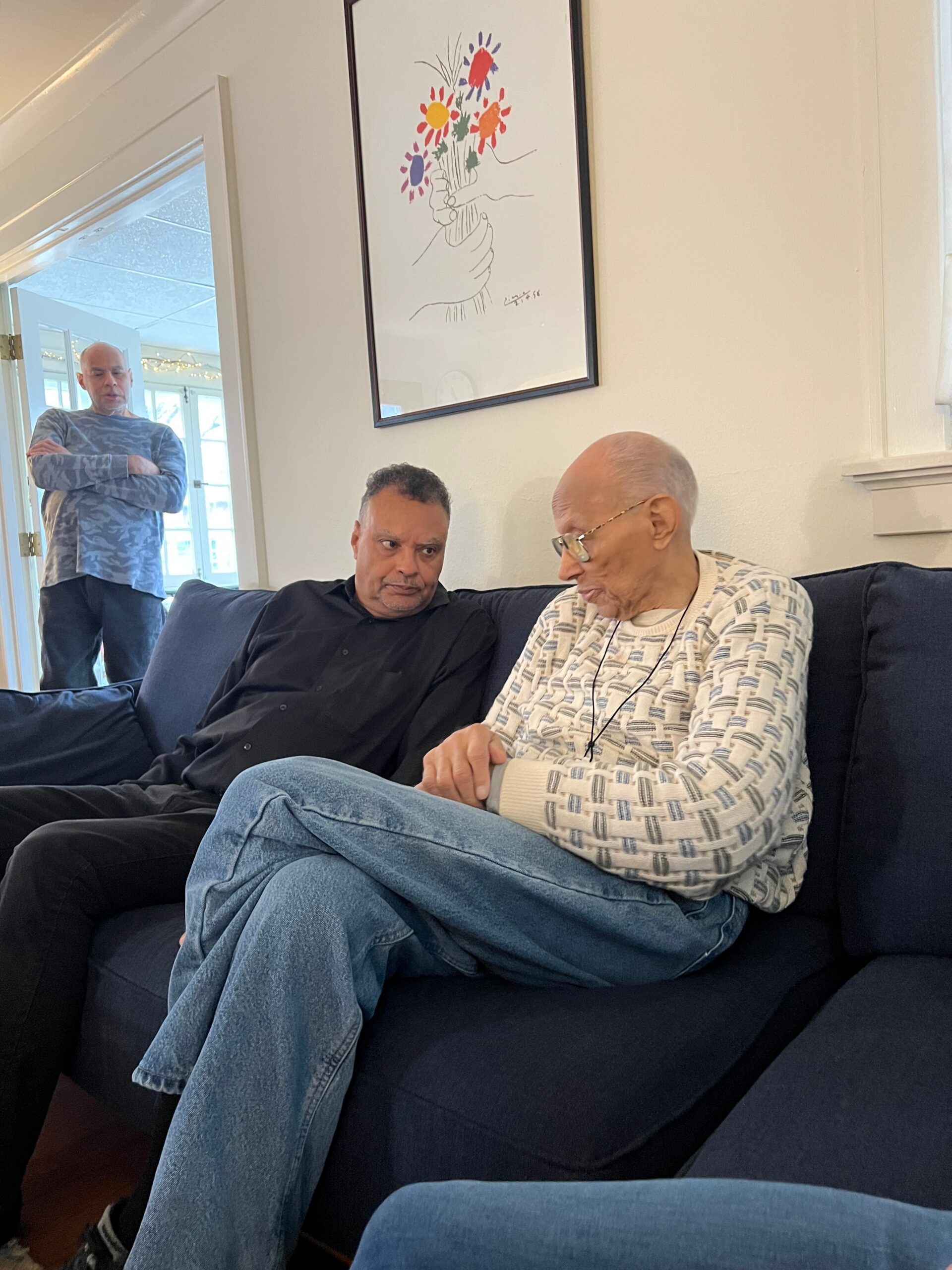 Three men in a living room: two seated on a dark sofa—one elderly with glasses, the other in a black shirt—while a third man stands near the doorway with arms crossed; a colorful bouquet artwork is on the wall behind them.