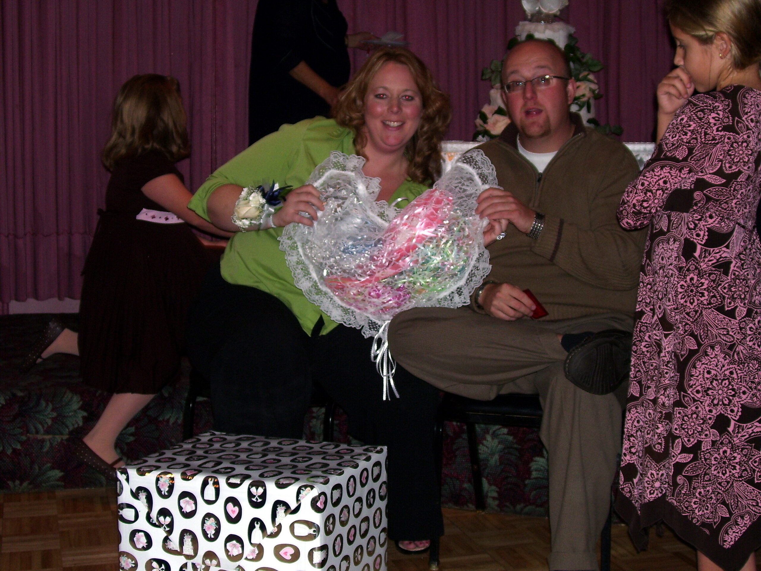 Smiling woman in a lime blouse holds a large lace-wrapped gift basket, with a man beside her, on a stage with purple curtains in the background.