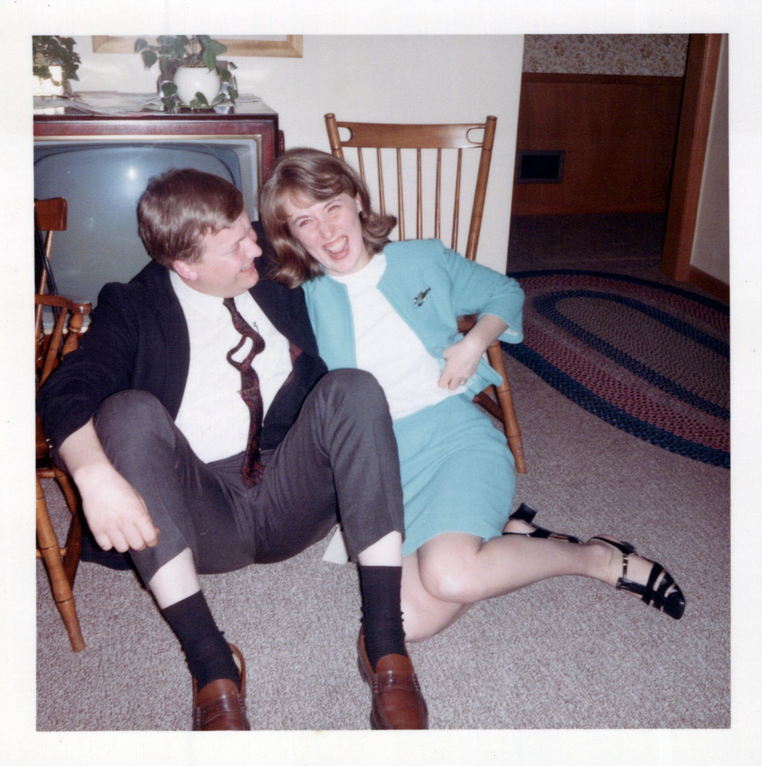 Two people sit on the floor indoors, smiling at the camera with wooden chairs nearby.